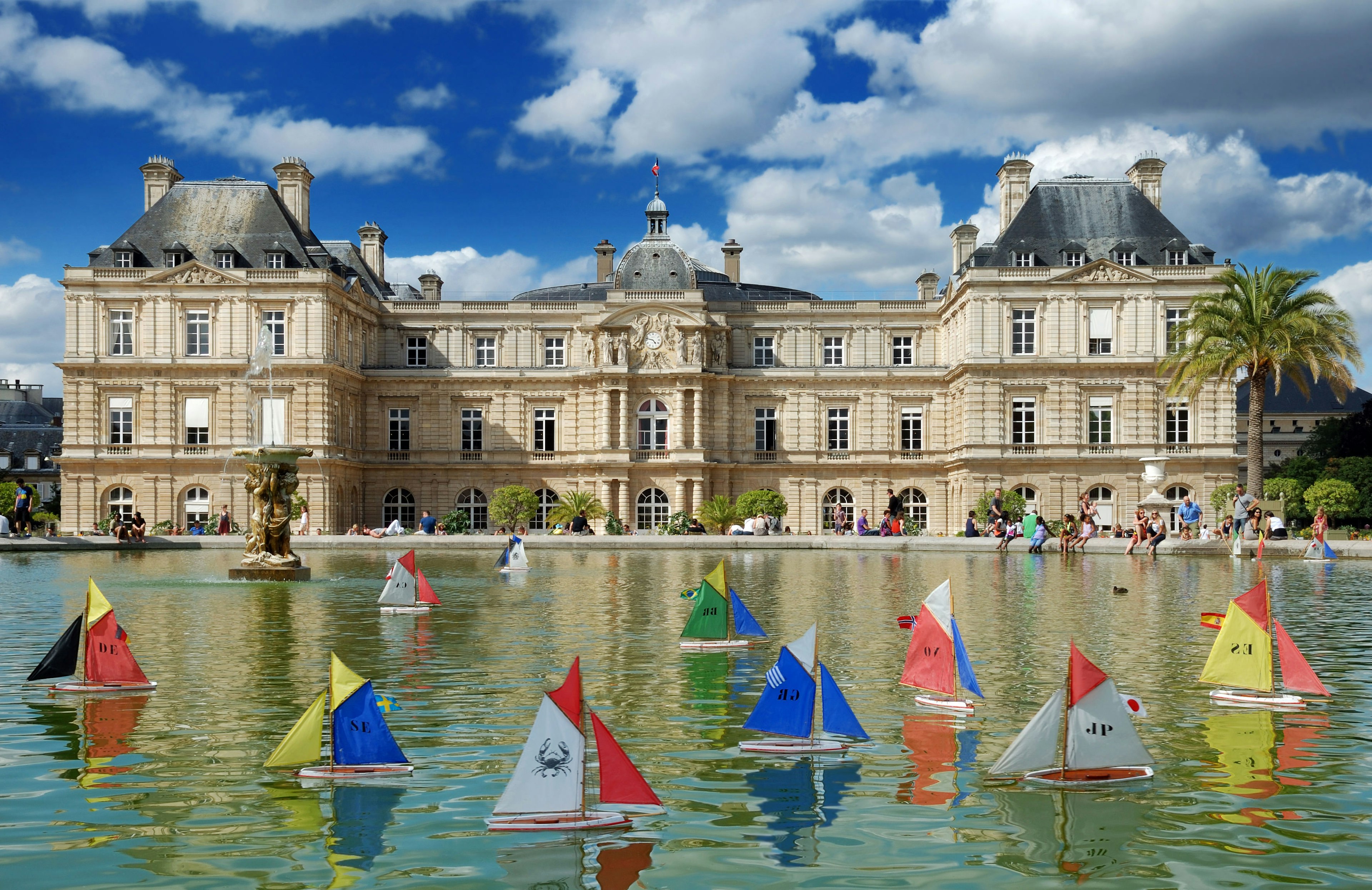 Colourful wooden toy boats sail on the still waters of the pond in Jardin du Luxembourg, Paris. There is a stately building with beige bricks in the background underneath a clear blue sky.