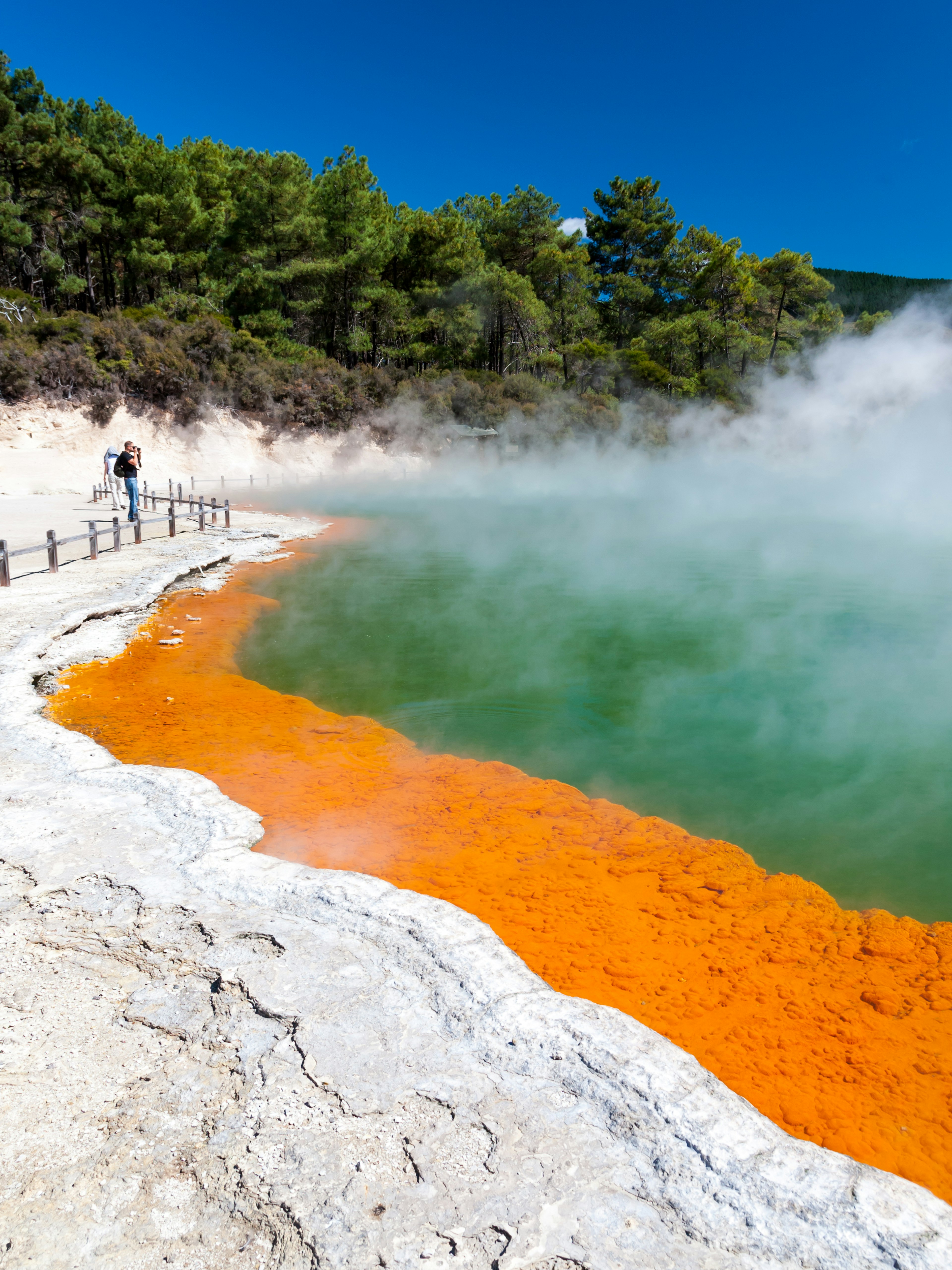 A figure stands on a walkway near a steaming hot spring