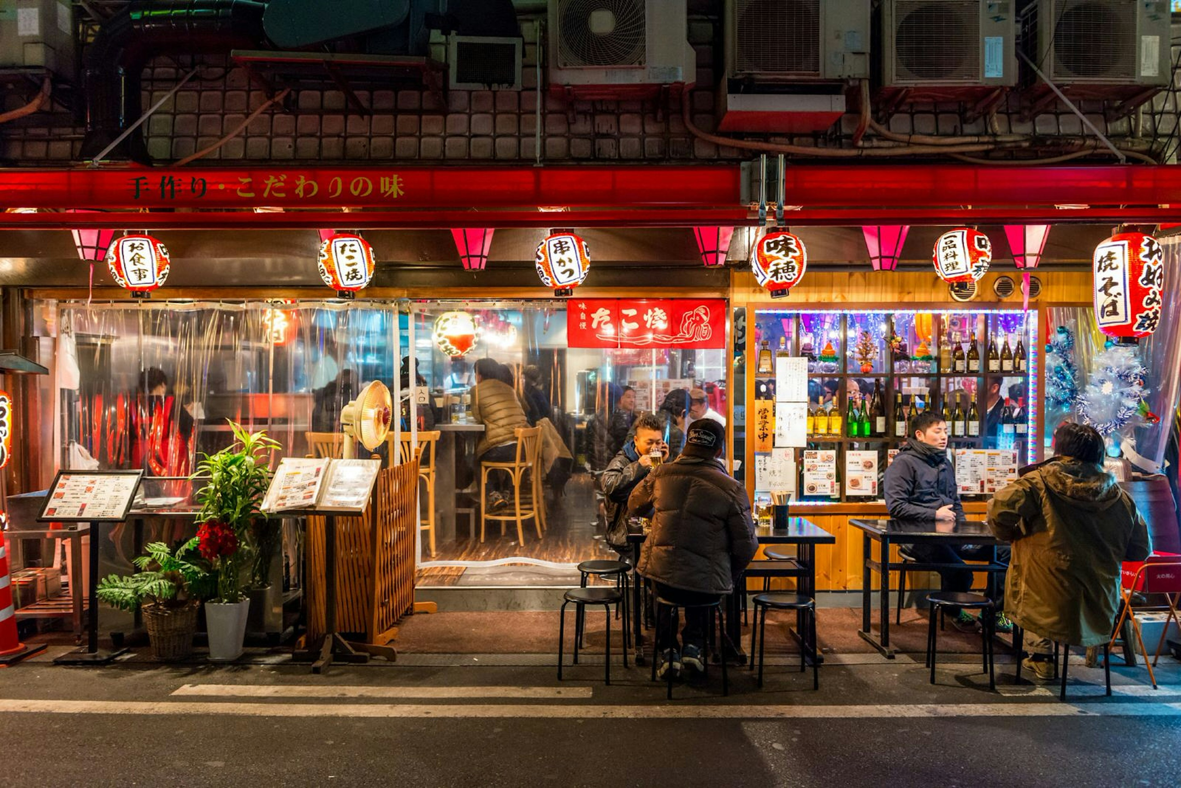 Locals gather at a restaurant in Osaka after dark. The customers are using outdoor seating, lit by red hanging lanterns. The windows are lined with bottles of alcohol and there are menus on display.