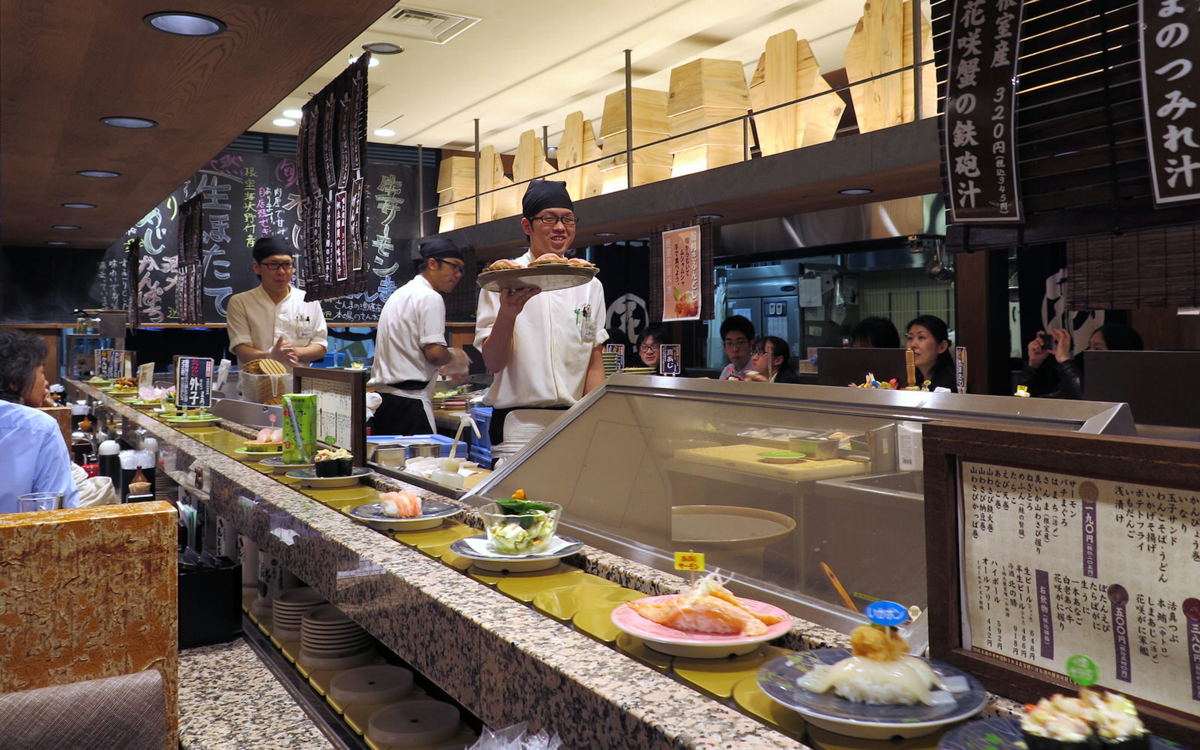 A Tokyo restaurant interior with a conveyor belt, upon which are platefuls of sushi and assorted Japanese fare. Behind the belt in the kitchen, chefs clad in white, smiling, prepare and hold aloft dishes.