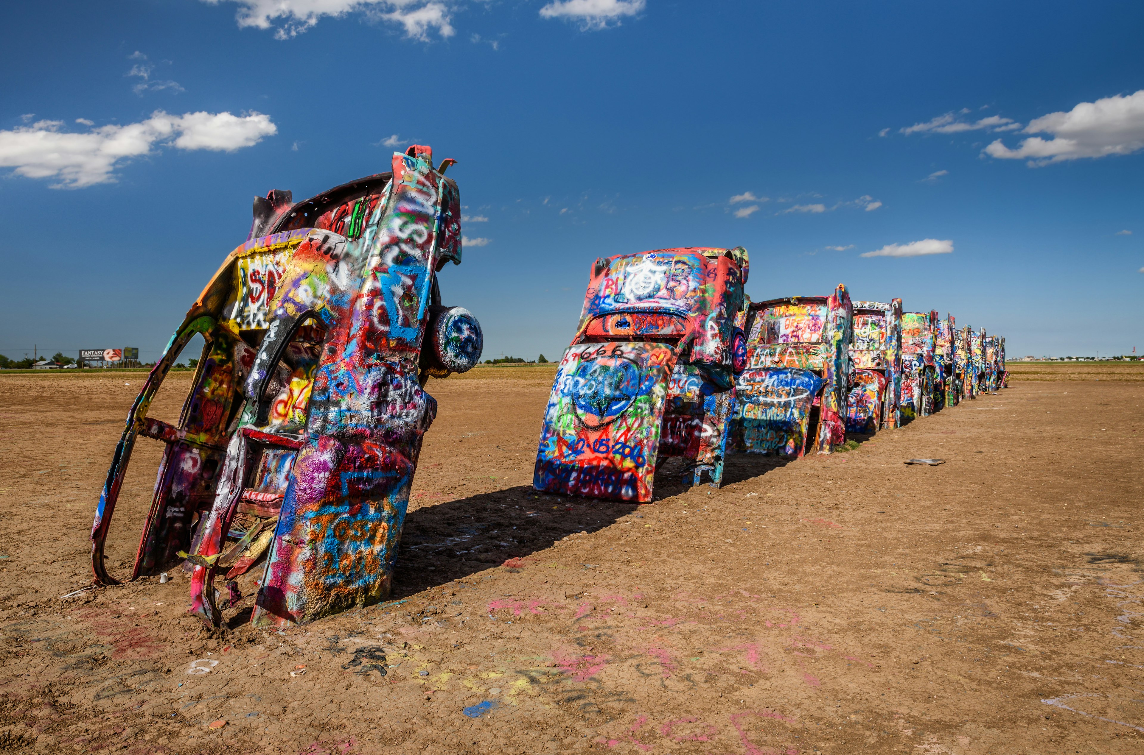 A row of 10 Cadillacs with their hoods buried in the earth stretches out. Each one is covered in colorful marks and graffiti