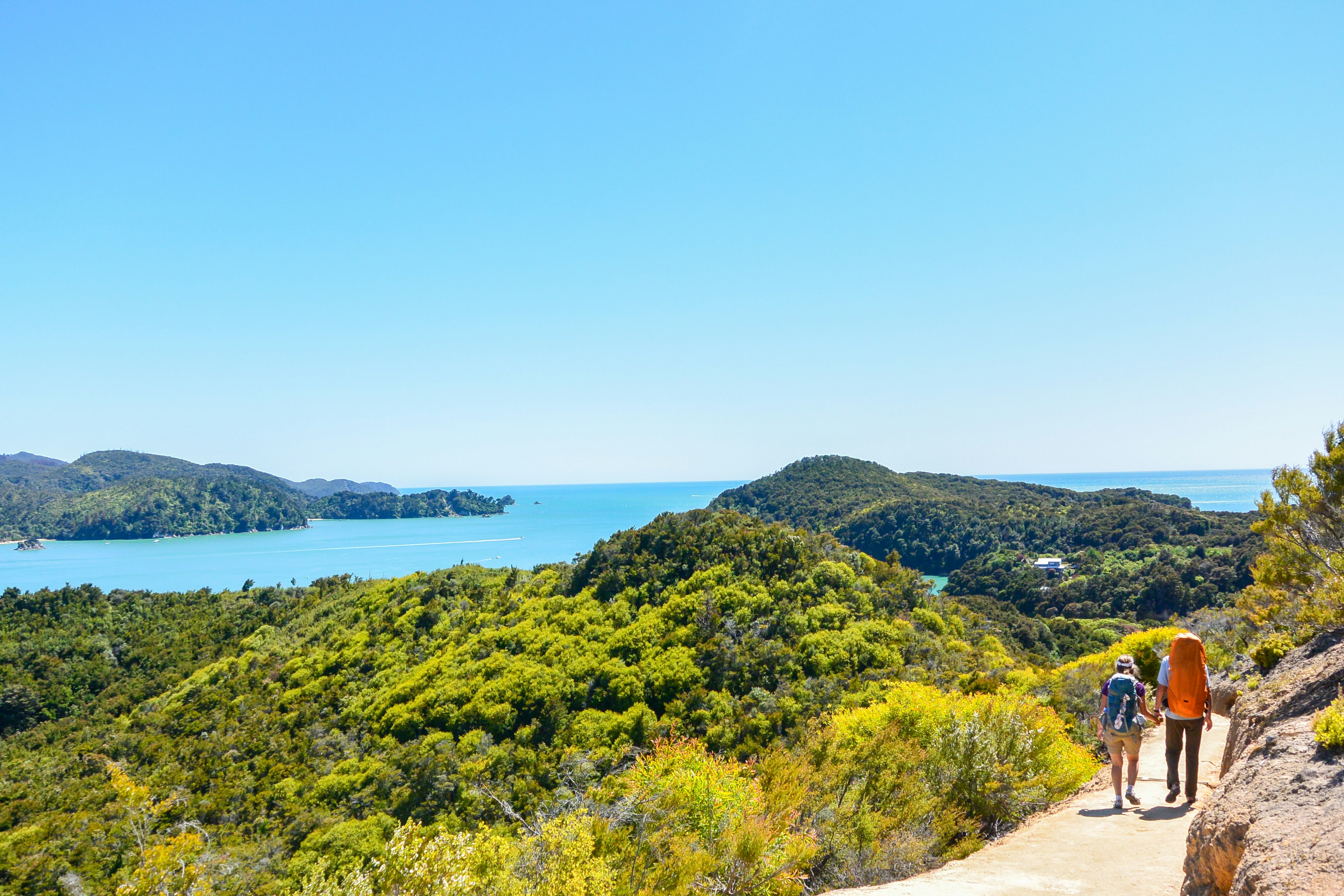 A couple walking with large backpacks along a coastal path; there is dense green scrub to the left of the path, with the bright blue sea beyond. To the right is a rocky hill.