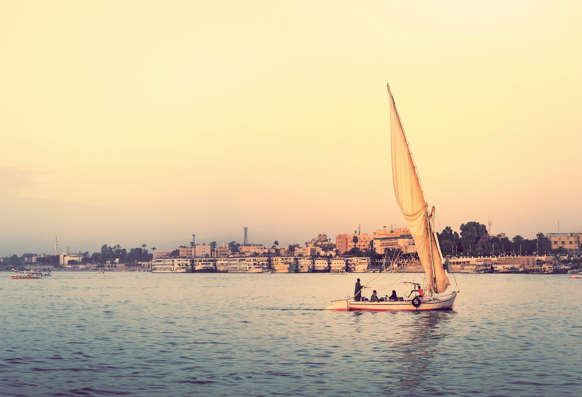 Felucca at sunset - travel on sail vessel on the Nile river, romantic cruise and adventure in Egypt. Traditional egyptian sailboat on horizon. Skyline of Luxor on riverside.