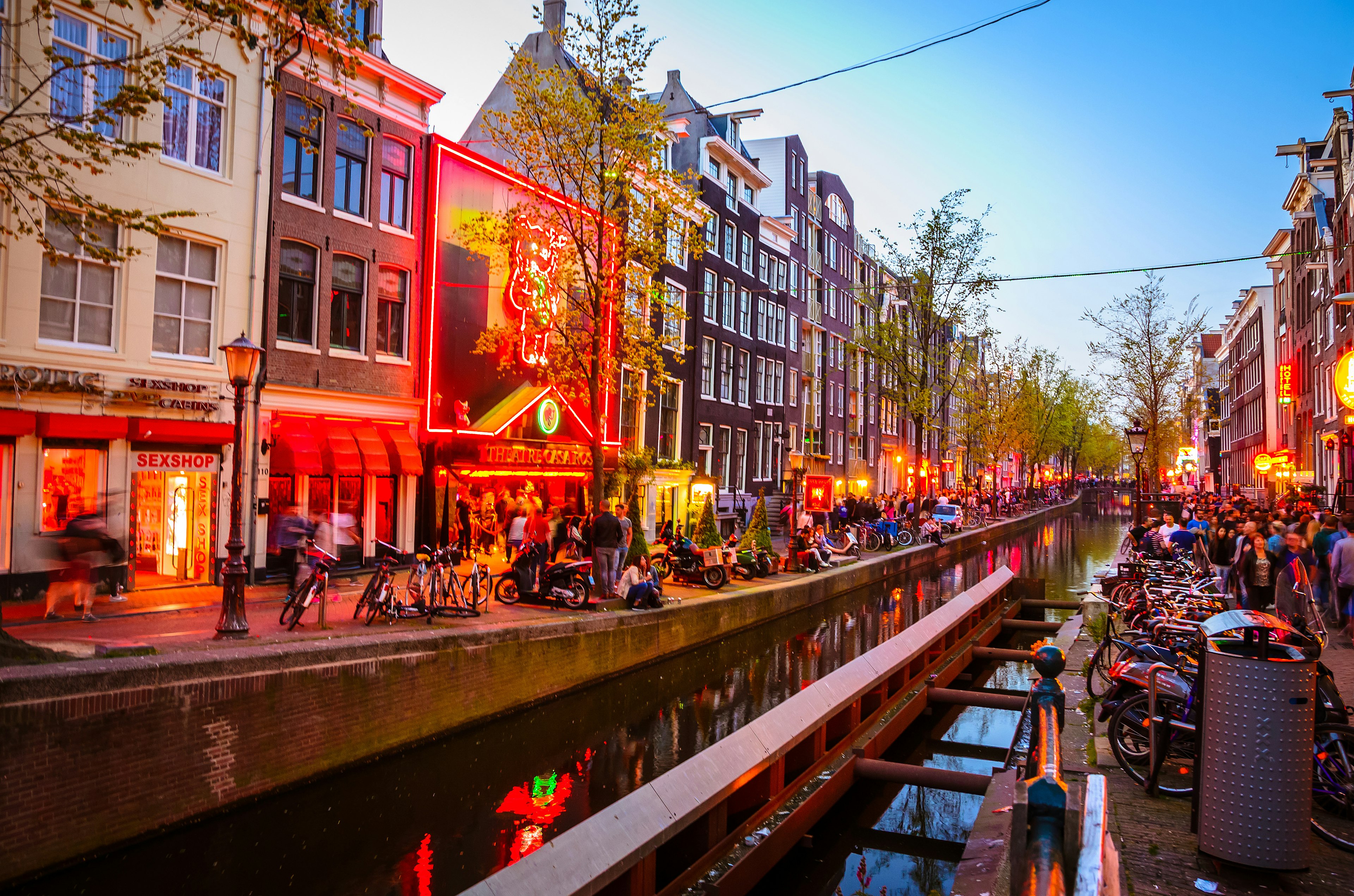 A canal cuts through the centre of Amsterdam's Red Light District, with bars and restaurants (lit in the iconic red lights) flanking the streets either side, while the roads are busy with people.