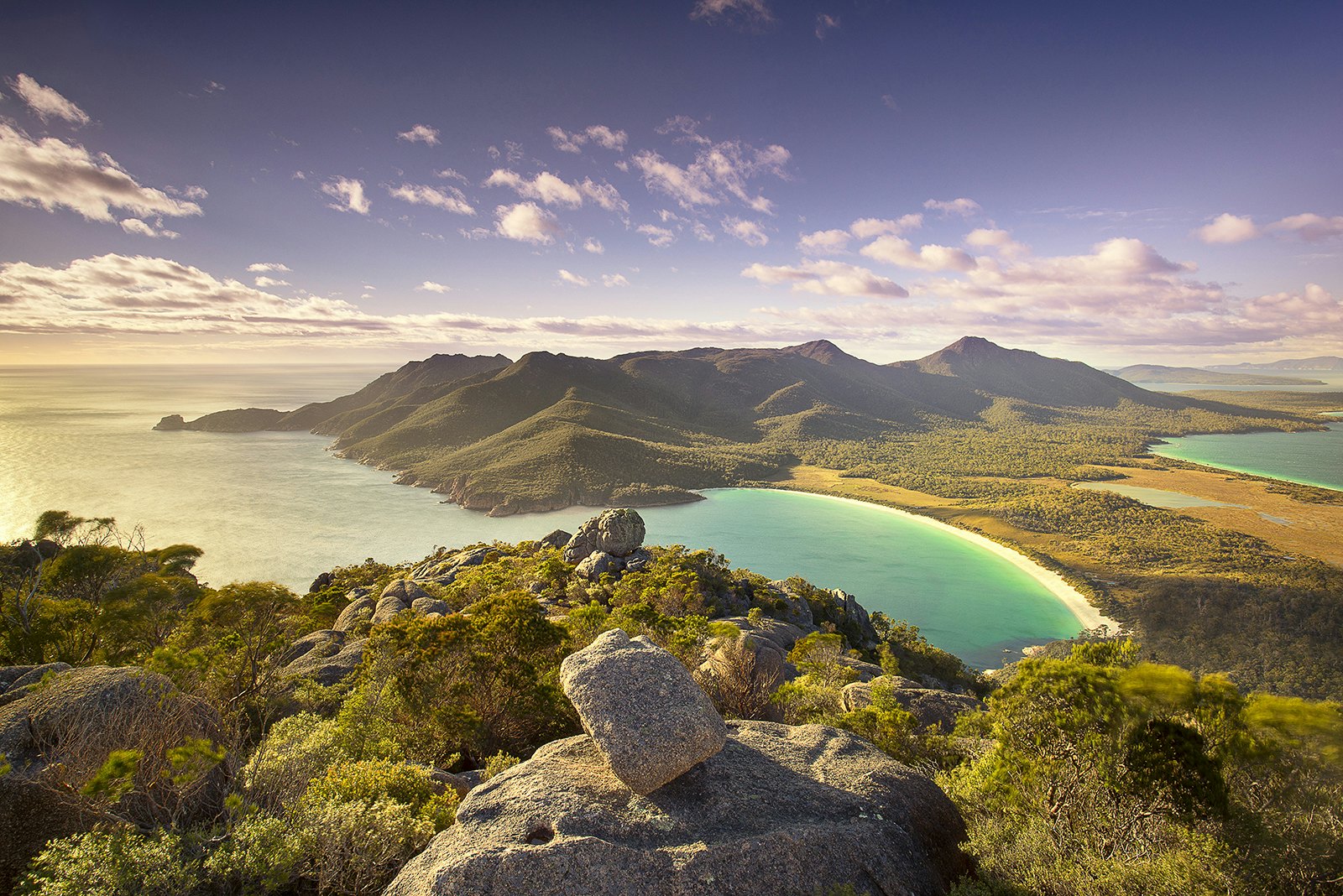 Top of Mt Amos over looking Wineglass Bay, Tasmania at dusk. There are rocks and shrubs in the foreground before giving away to a beautifully smooth beach and blue sea. The sun is low in the sky, throwing out very ambient light.