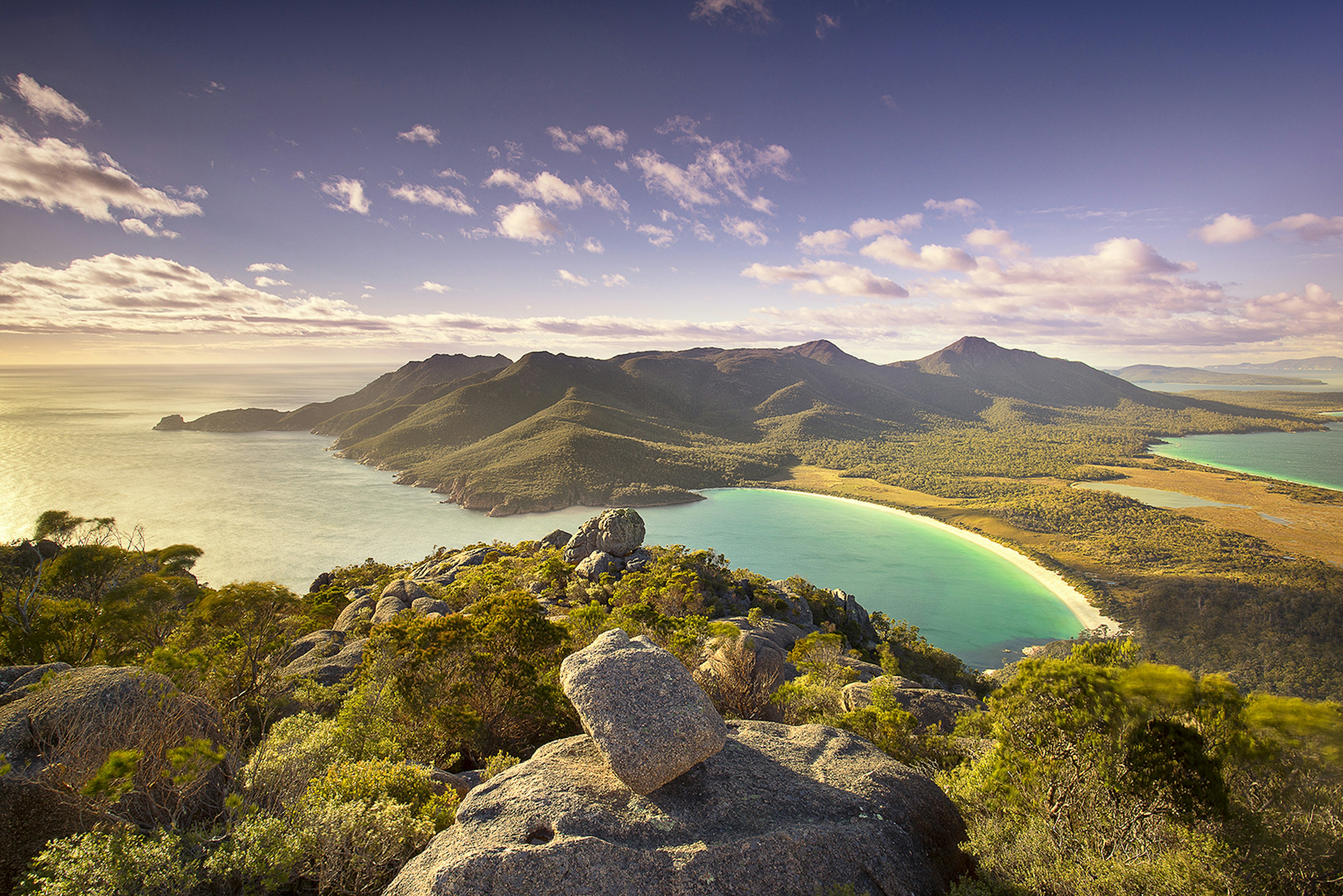 Top of Mt Amos over looking Wineglass Bay, Tasmania at dusk. There are rocks and shrubs in the foreground before giving away to a beautifully smooth beach and blue sea. The sun is low in the sky, throwing out very ambient light.