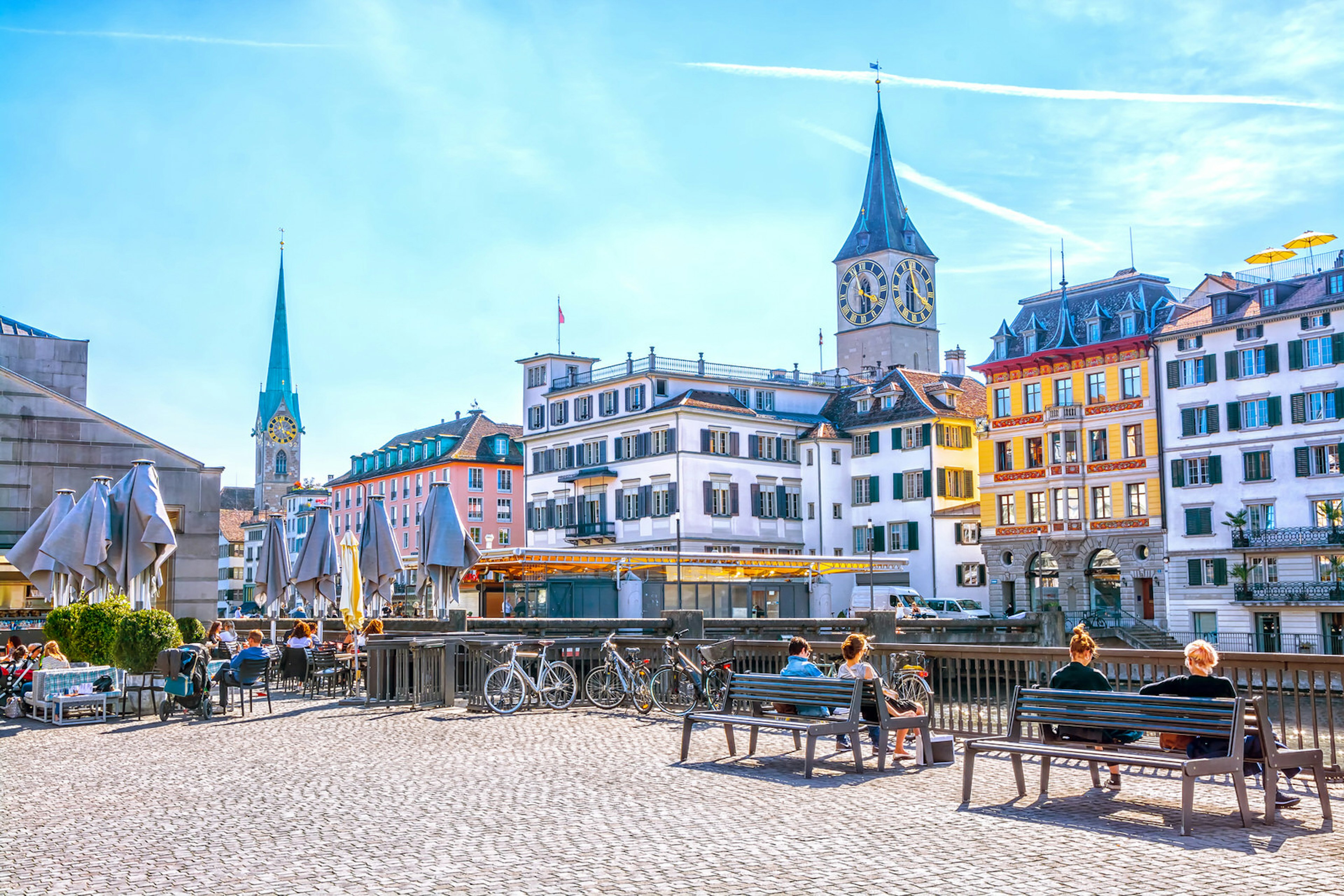 People relax on benches on Limmat River quay in Zurich, with the Fraumunster church in the background