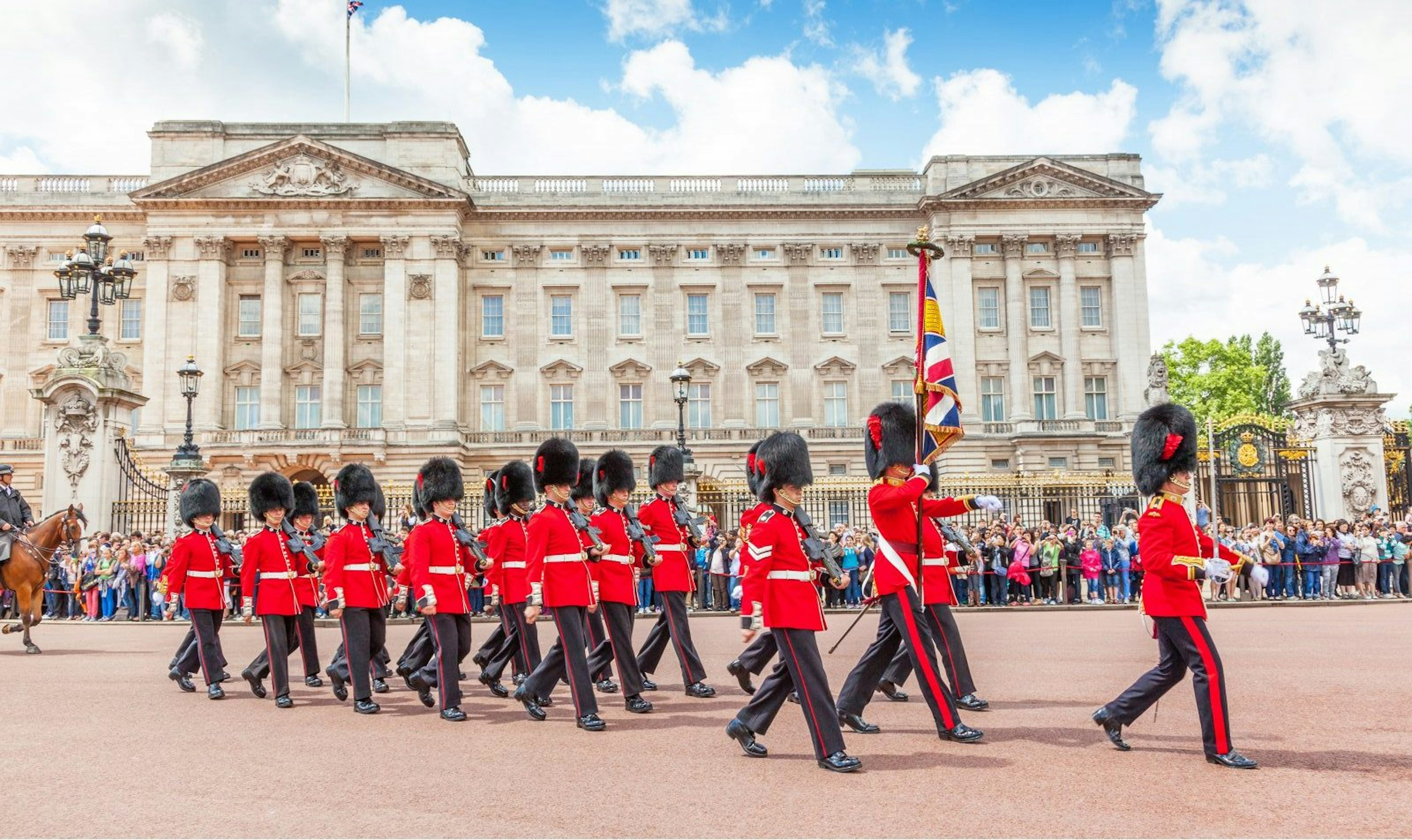 Guards in red jackets an bearskin hats march in front of Buckingham Palace