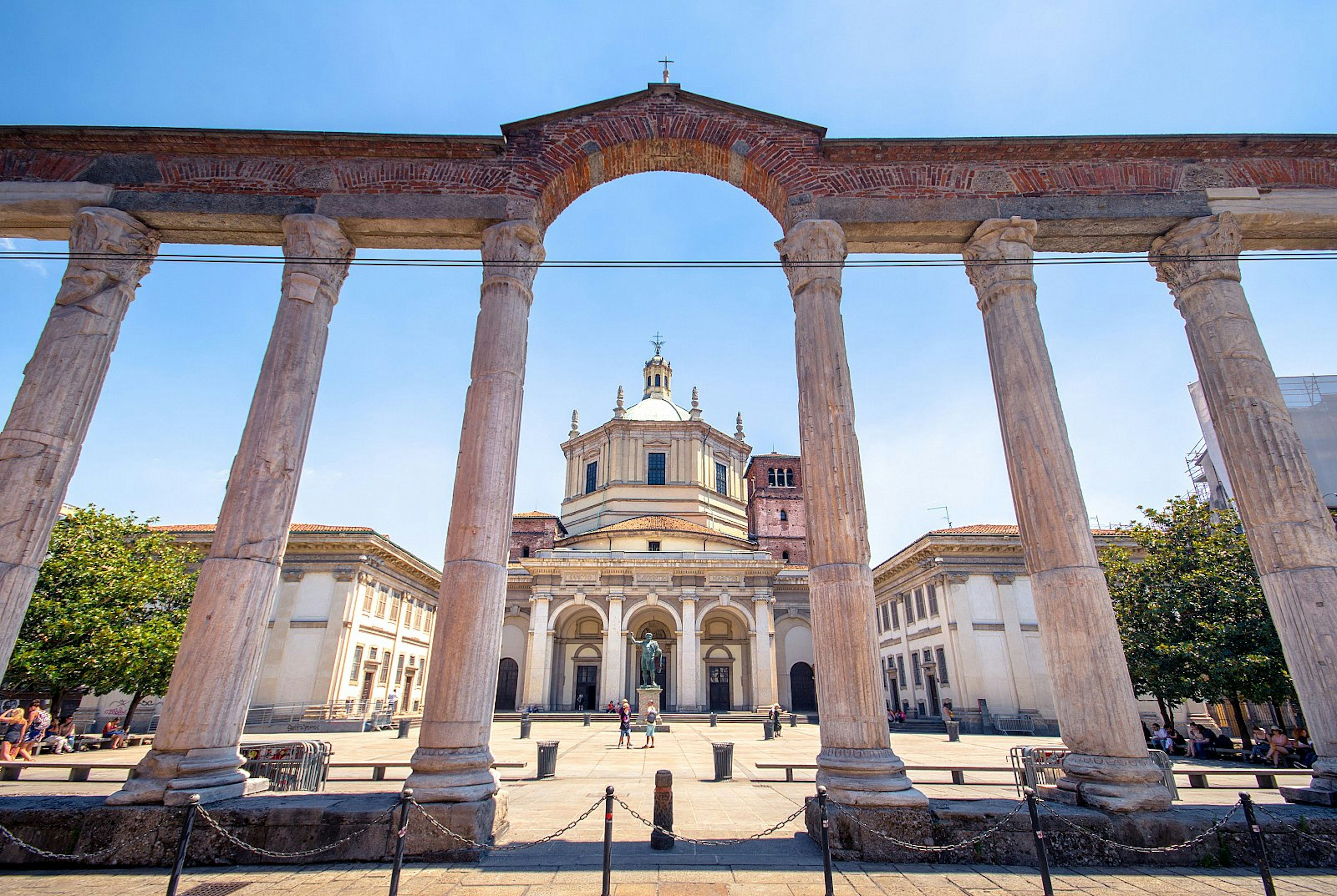 The San Lorenzo Columns, a row of stone Roman-era columns with a tree-lined piazza and a church beyond.