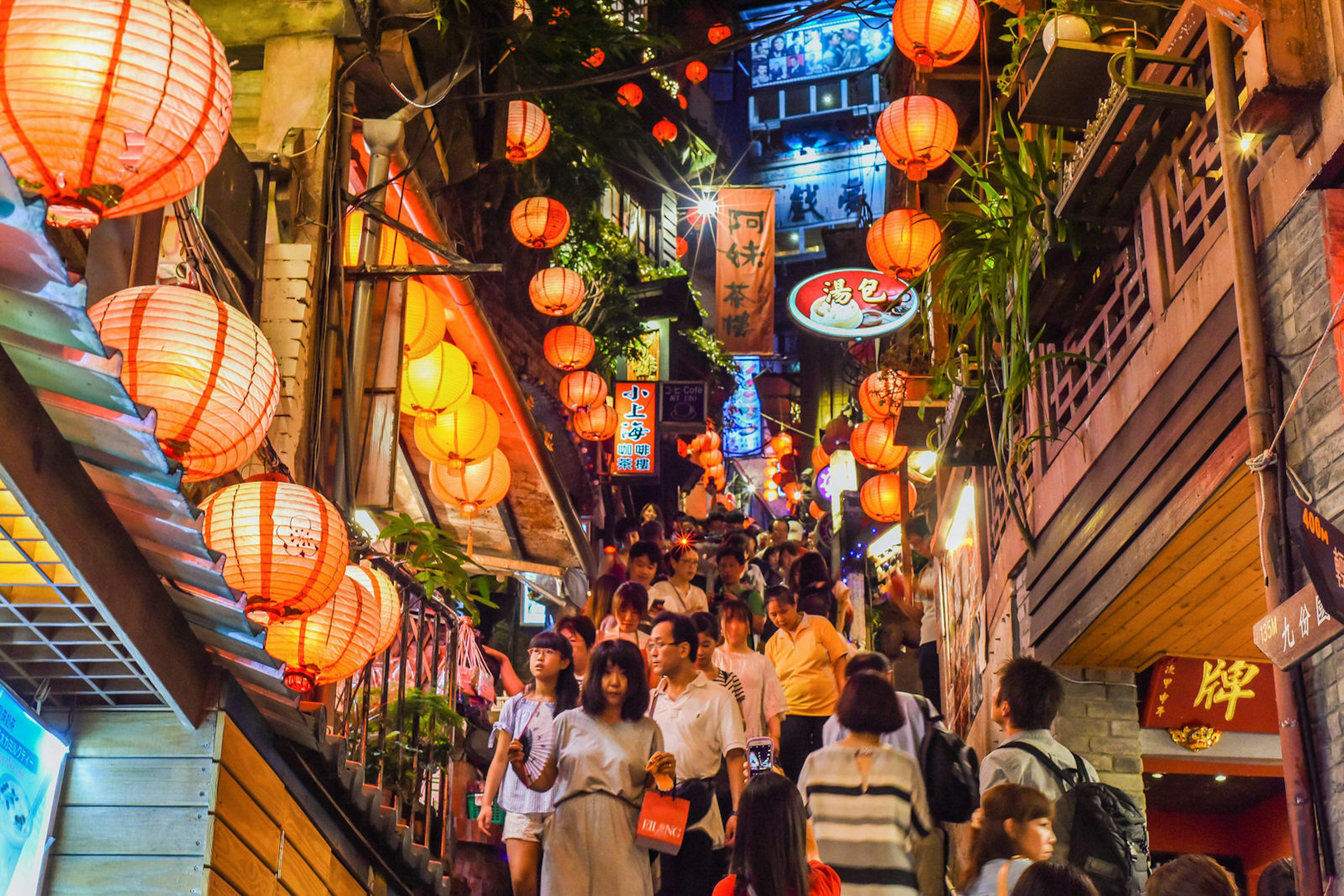 A throng of people are travelling up and down a steep street in Taiwan at night; the street is strung with orange lanterns, while wooden buildings are arrayed with vivid Taiwanese signs and greenery.