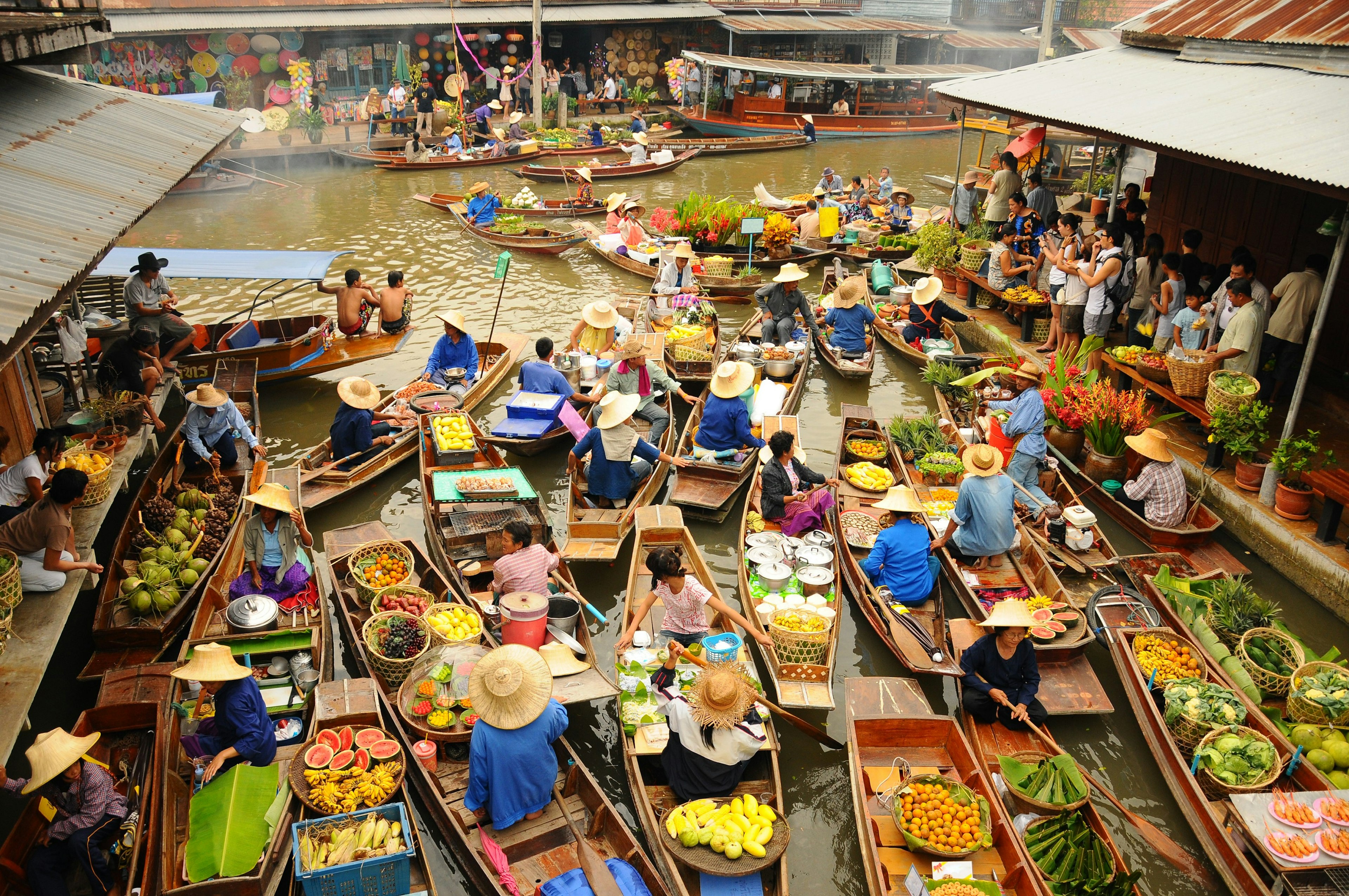 A vast amounts of small wooden boats gather to trade goods at  Amphawa floating market. Most of the boats sell food, specifically fruits. Tourist wielding cameras watch on.