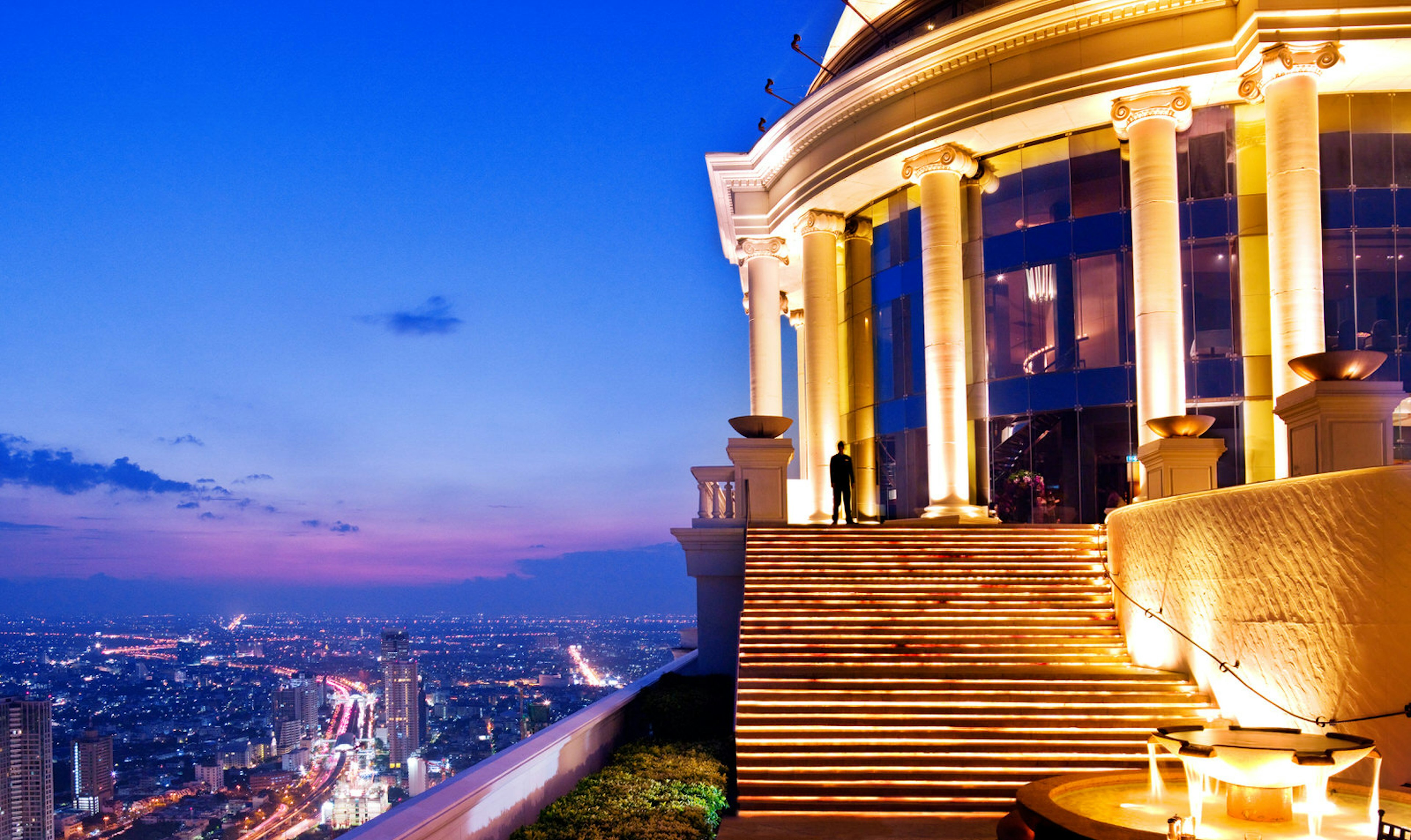 A Roman-looking circular stone building with large columns is lit up brilliantly from below; to the left, down in the distance is the teeming city of Bangkok