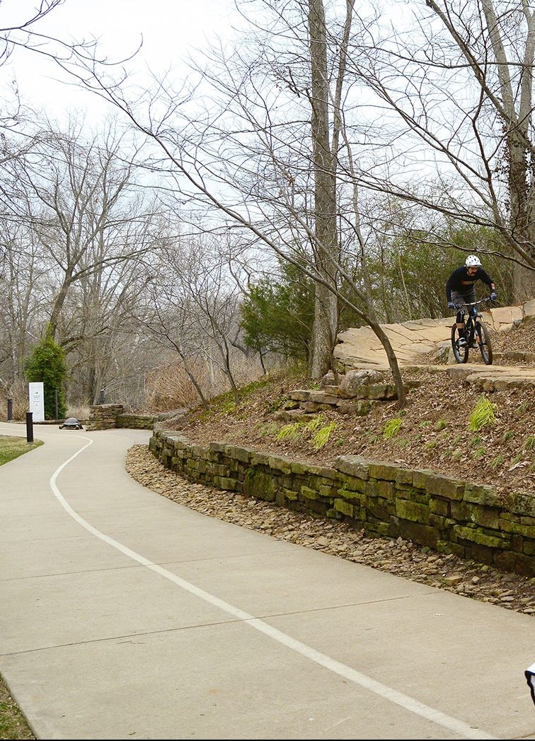 A man and a woman walk along a pedestrian path, with a mountain biker and a mountain bike trail in the background