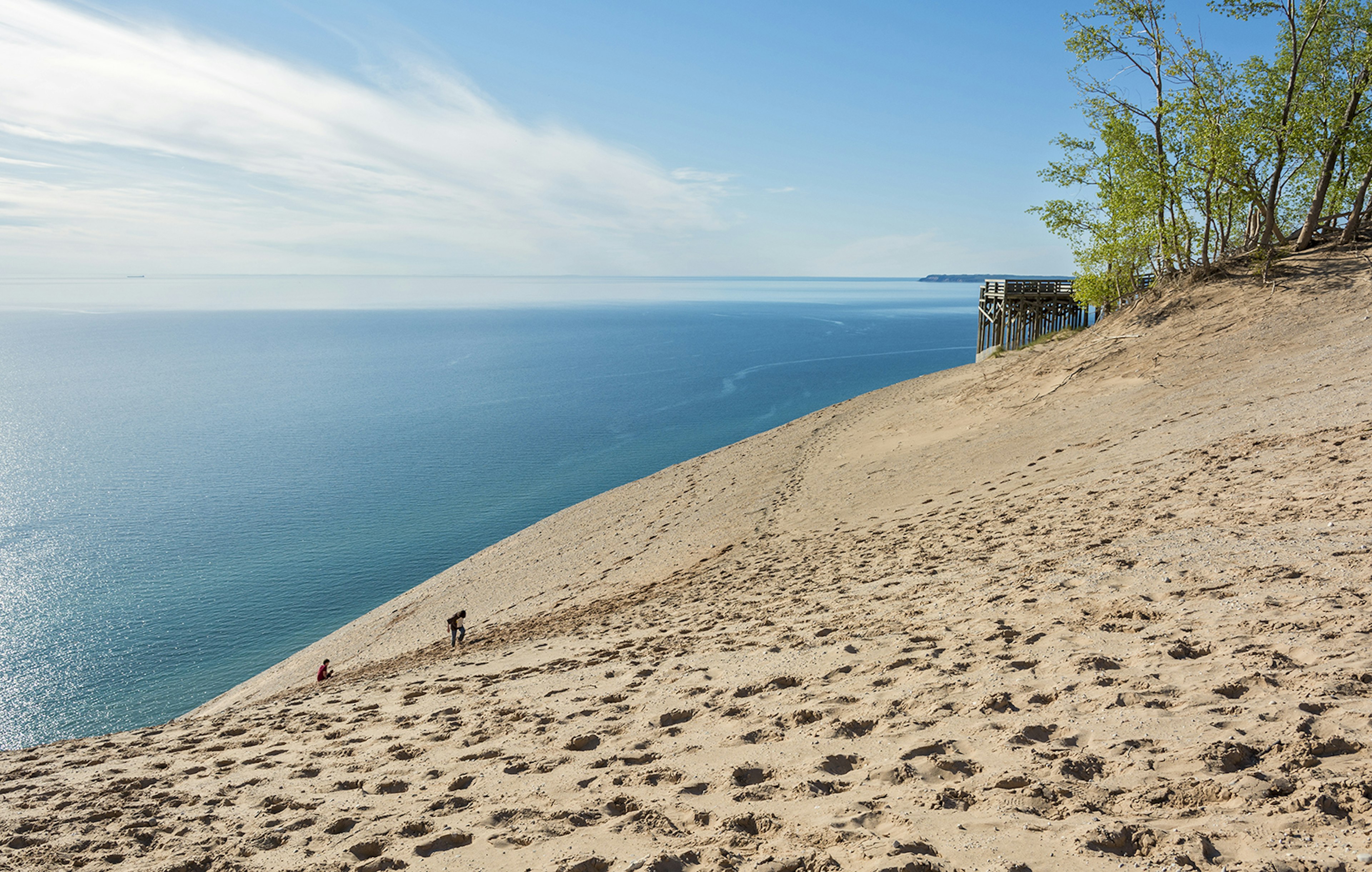 Two people hike up a massive golden sand dune with Lake Michigan in the background