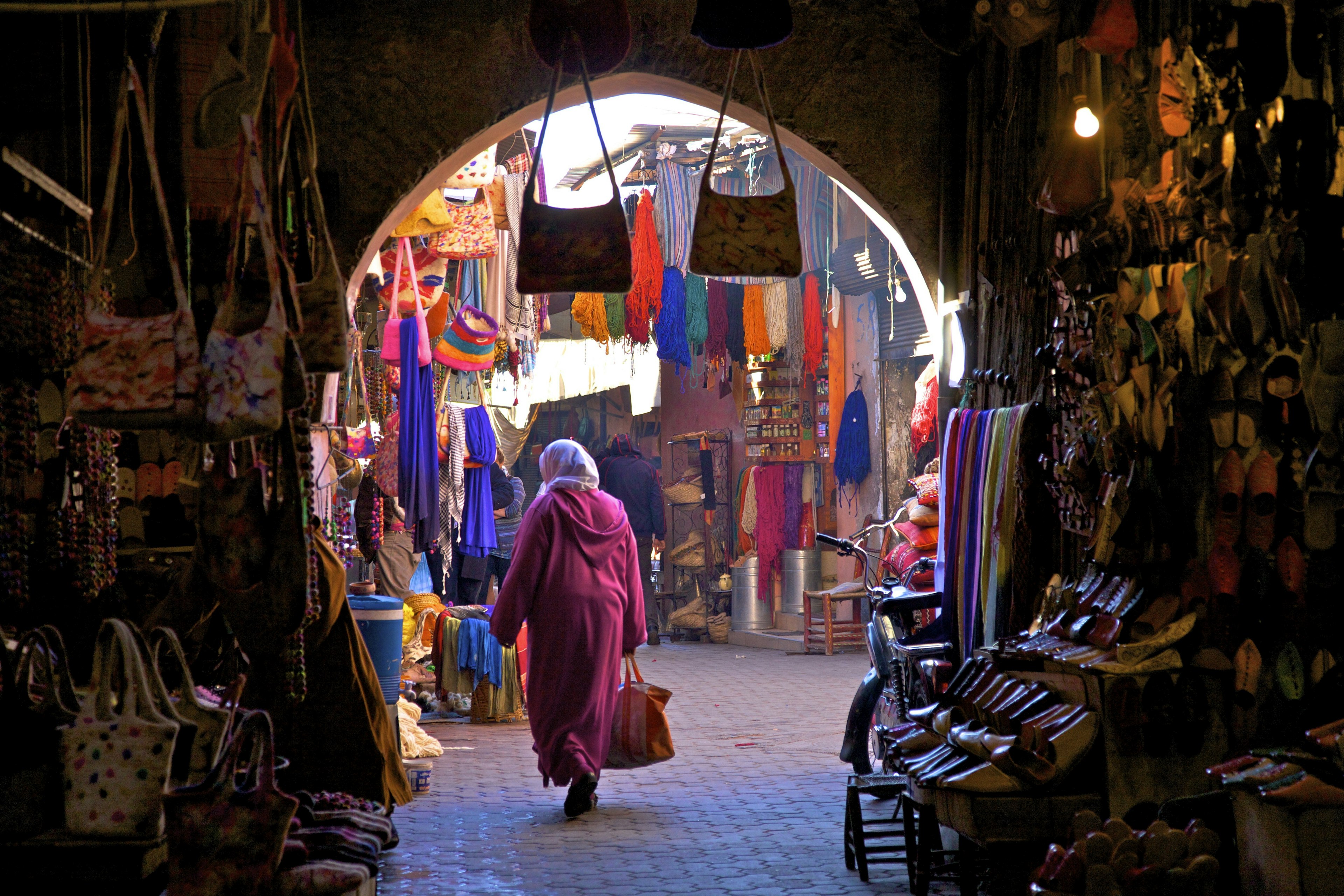 A woman in a pink dress and white hijab walks through one of the iconic archways of Morocco in a colorful souk in Marrakech