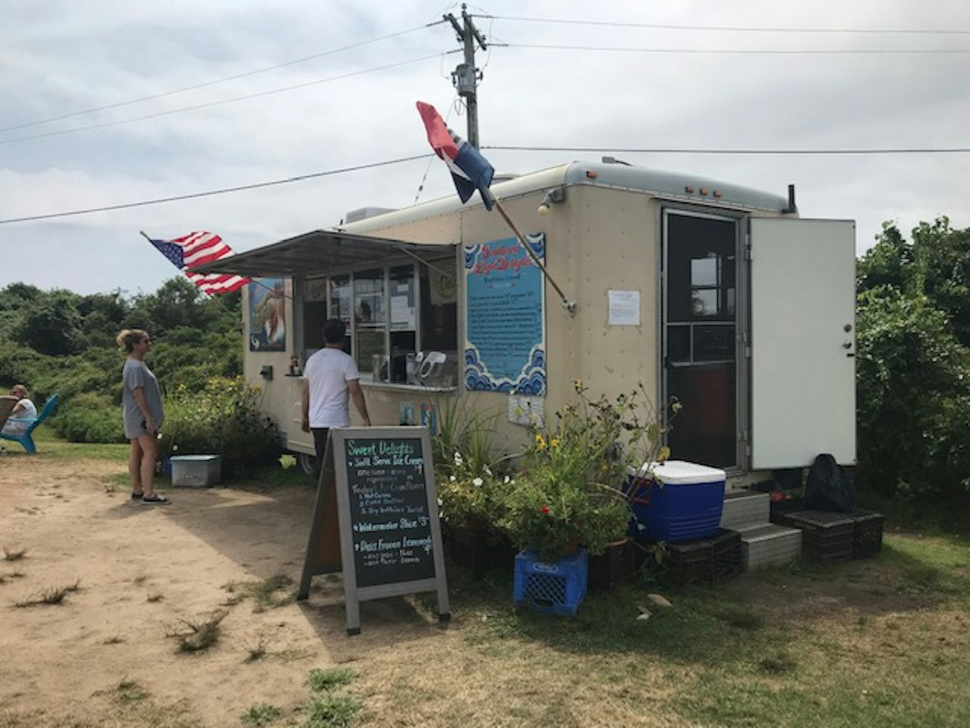 A food stall called 'Southeast Light Delights'. There's a man in a white t-shirt ordering food, and an American flag draped from the stall is blowing in the wind.