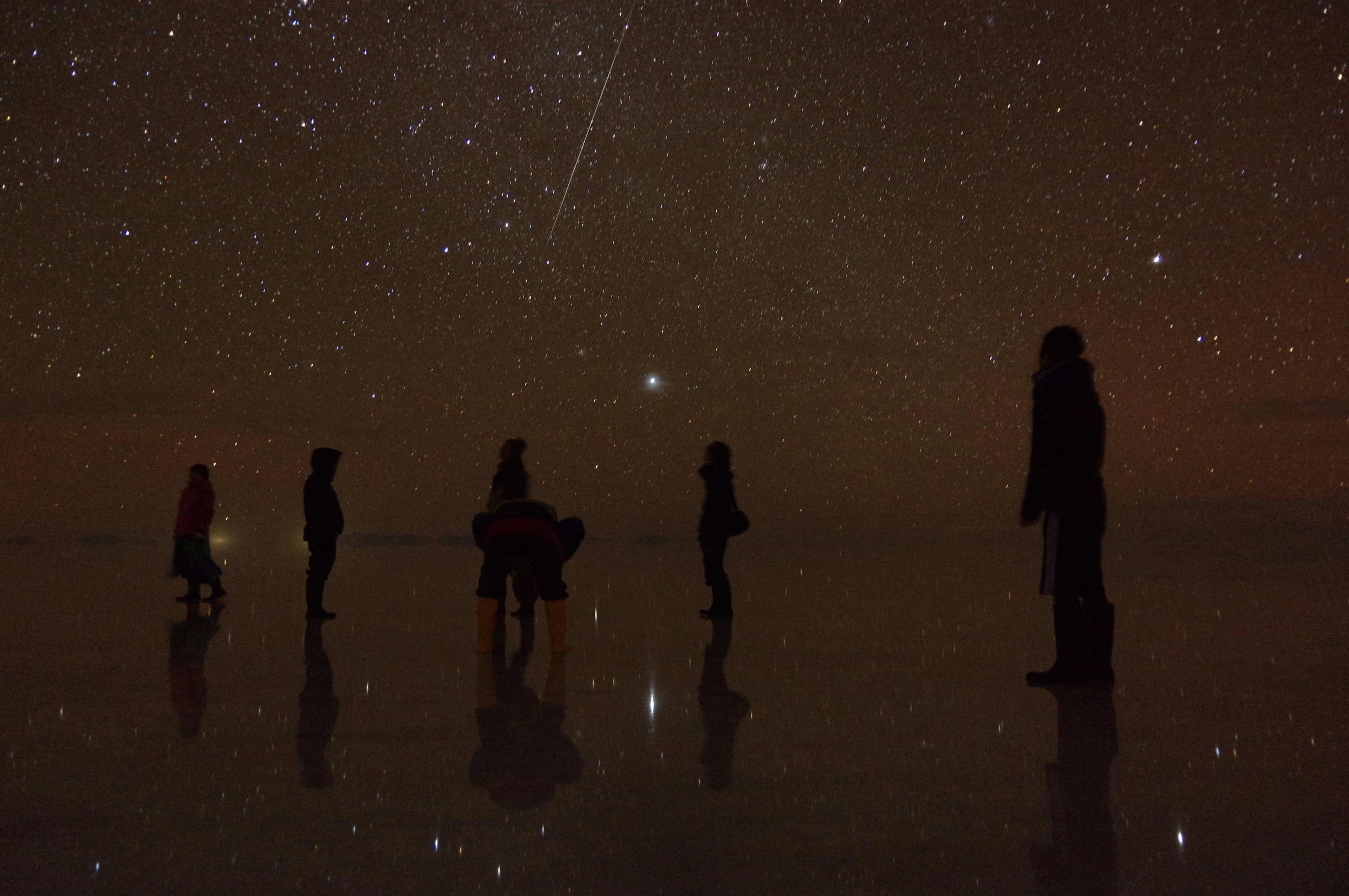 People stand on Bolovia's Salar de Uyuni at night; water covers the surface, so stars reflect off the ground, mirroring the sky. Bolivia, South America.