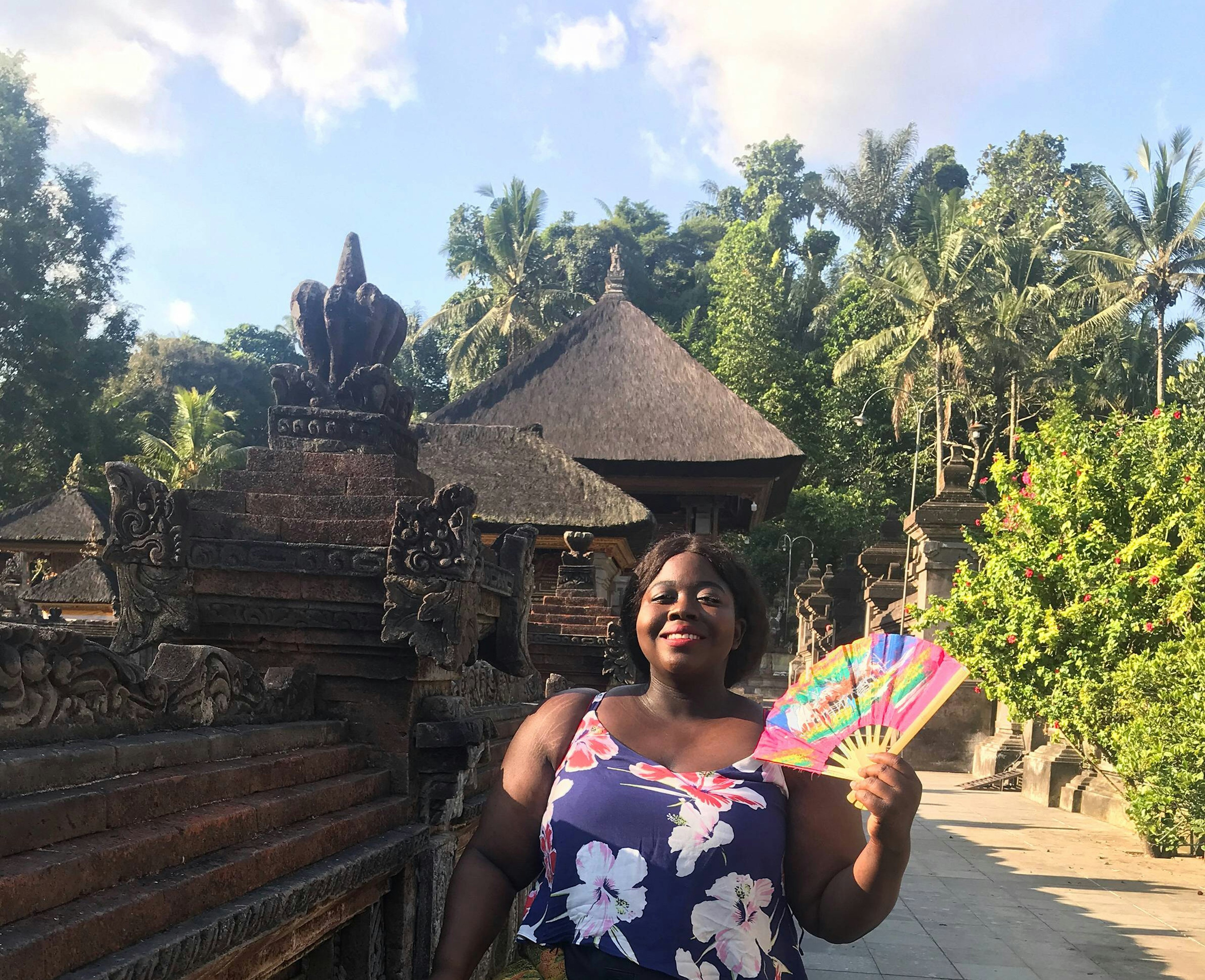 Stephanie Yeboah fanning herself while exploring a cultural site. Stephanie is wearing a floral-print top and holding a multi-coloured fan in front of statues and thatch-roofed buildings