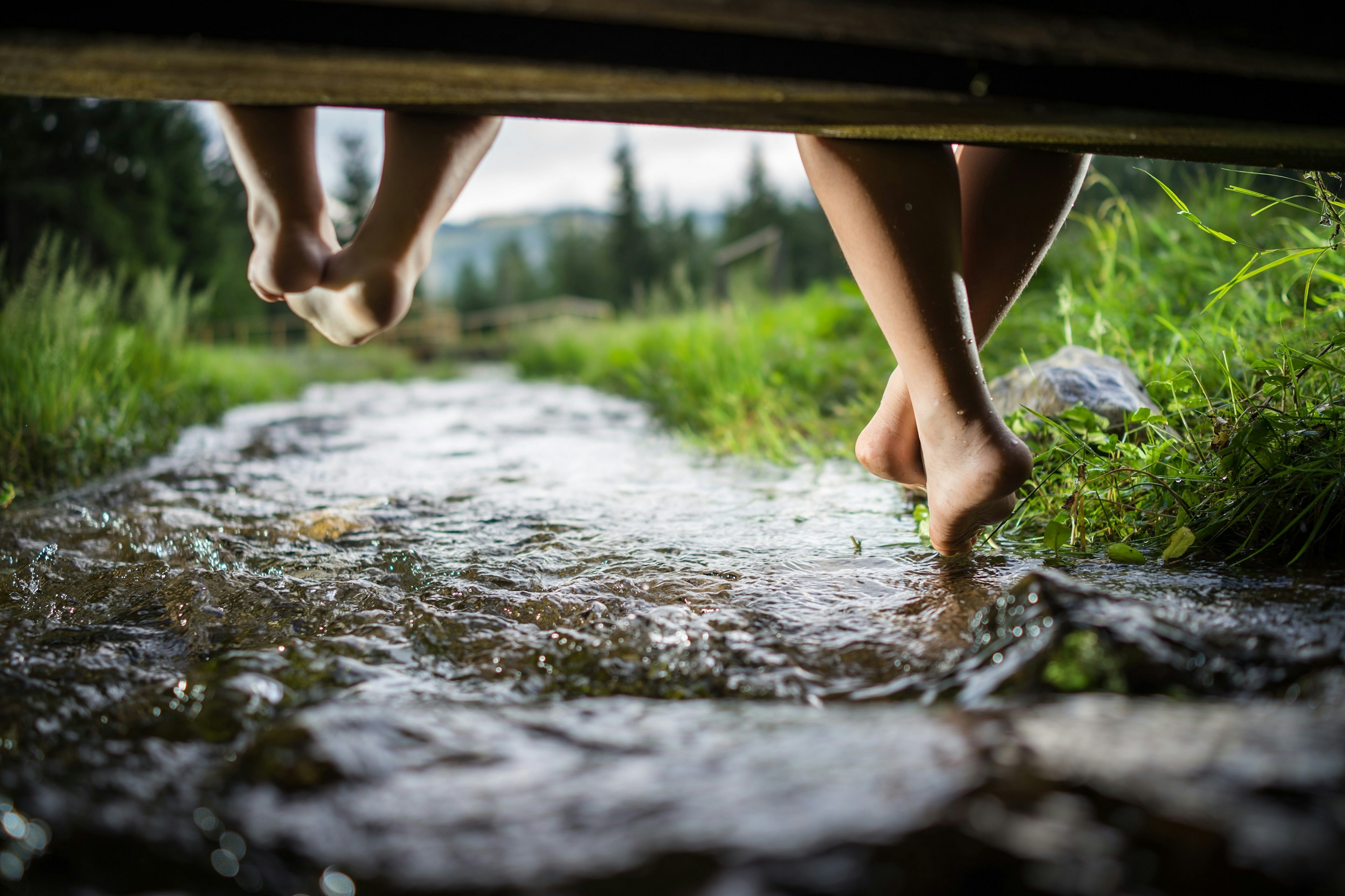 Children's feet dangling over a small stream from a wooden walkway in a mountainous area.