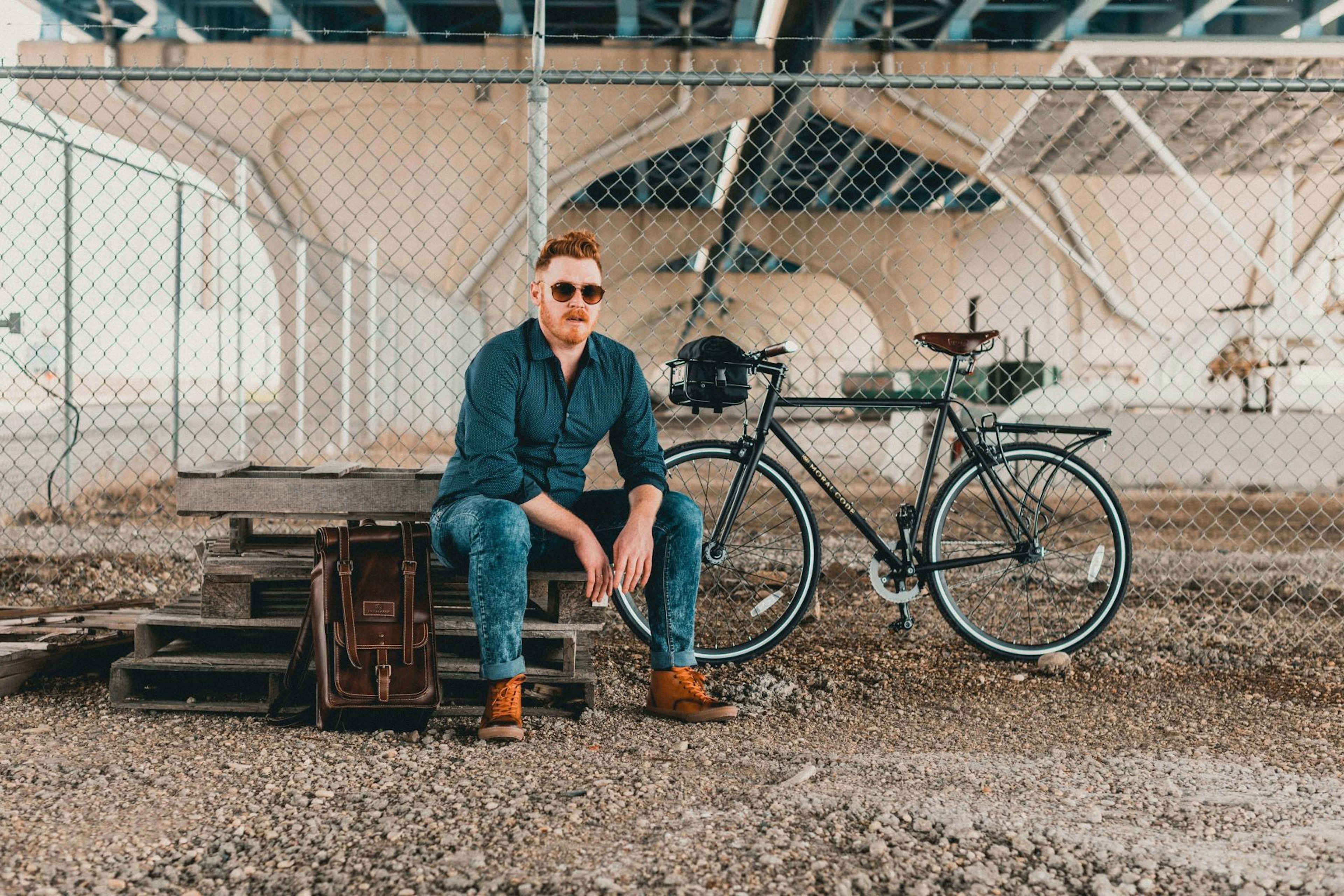 A bearded man sits on a pile of pallets with a bike and backpack nearby