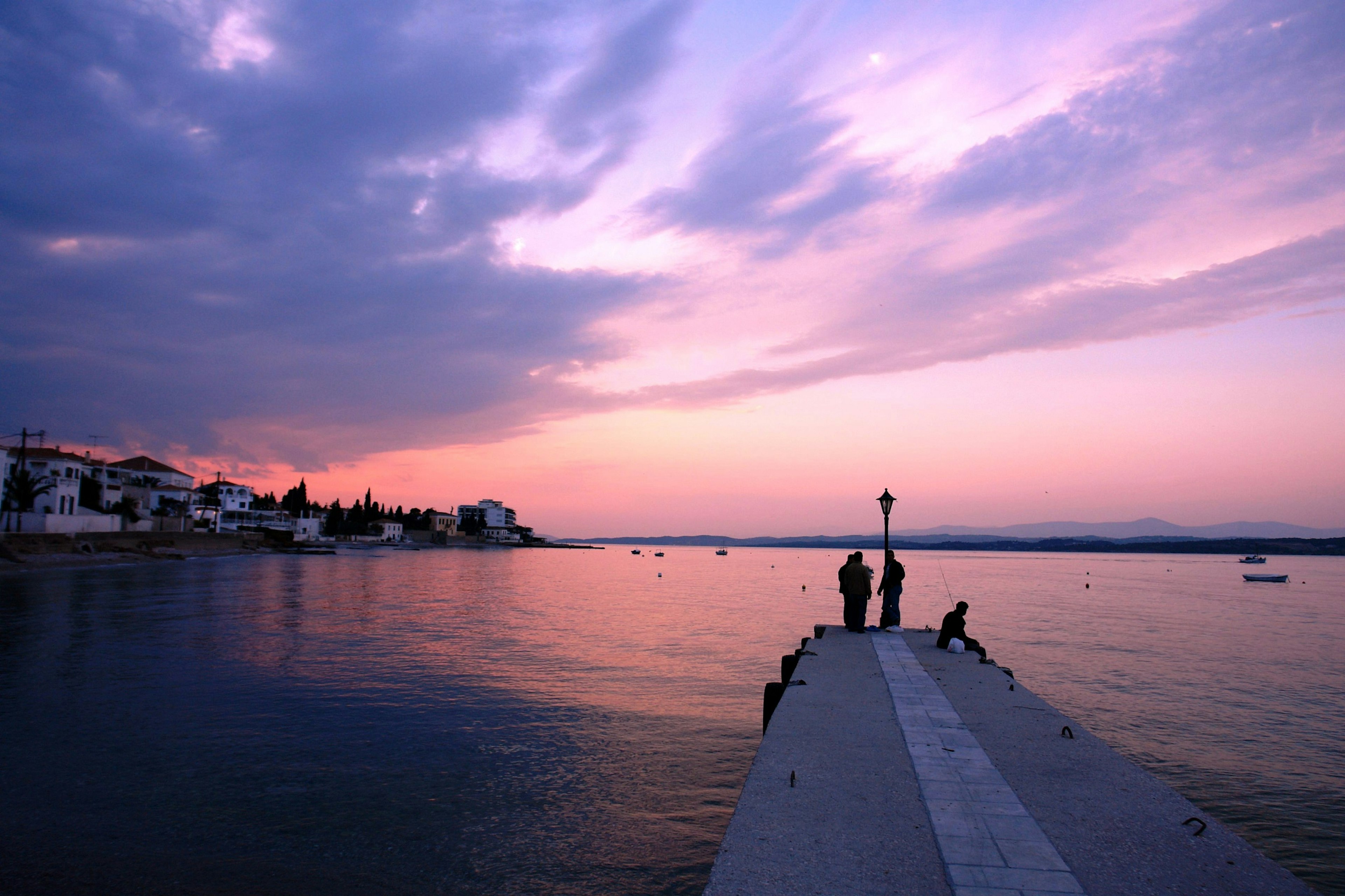 A pink sky over the sea seen from the shore of a Greek Island. A stone pier juts out into the sea with a lamppost at the end of it, with some people fishing at the end of the pier.