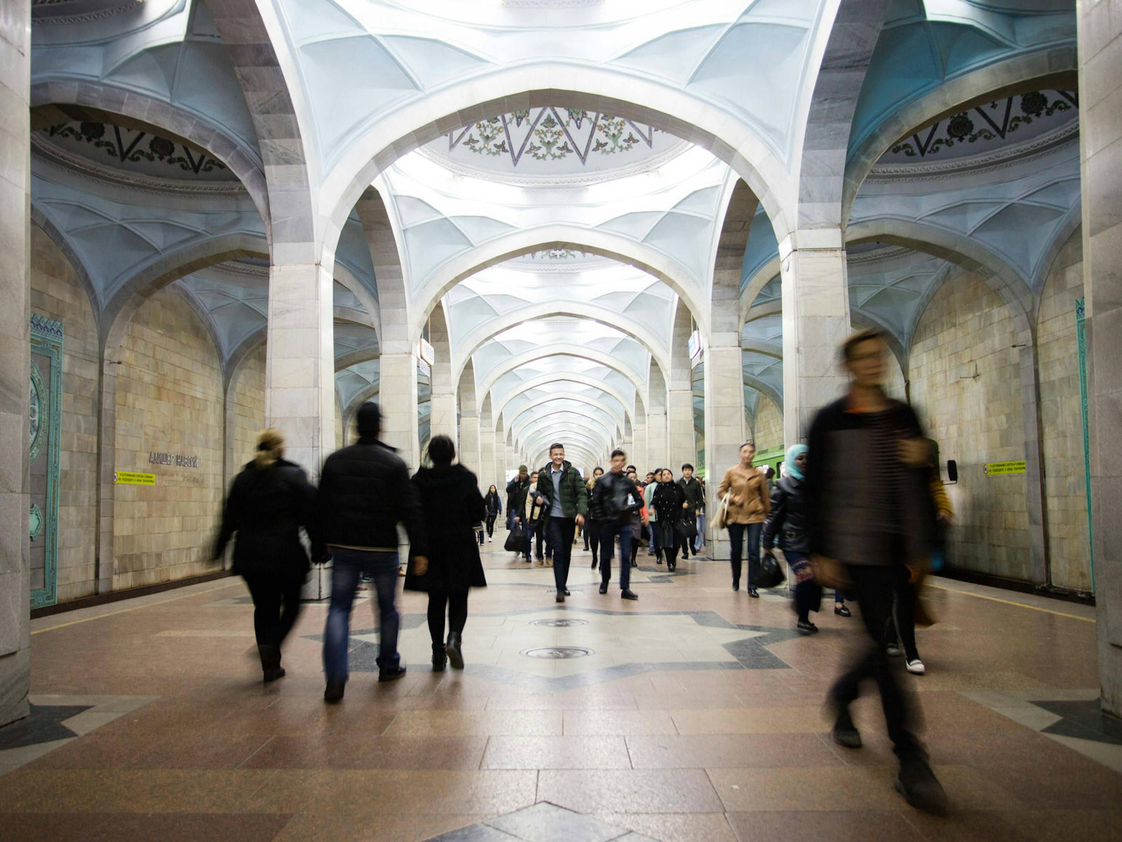 View of Alisher Navoi station with its blue arched domes and blurred passengers walking down the platform.