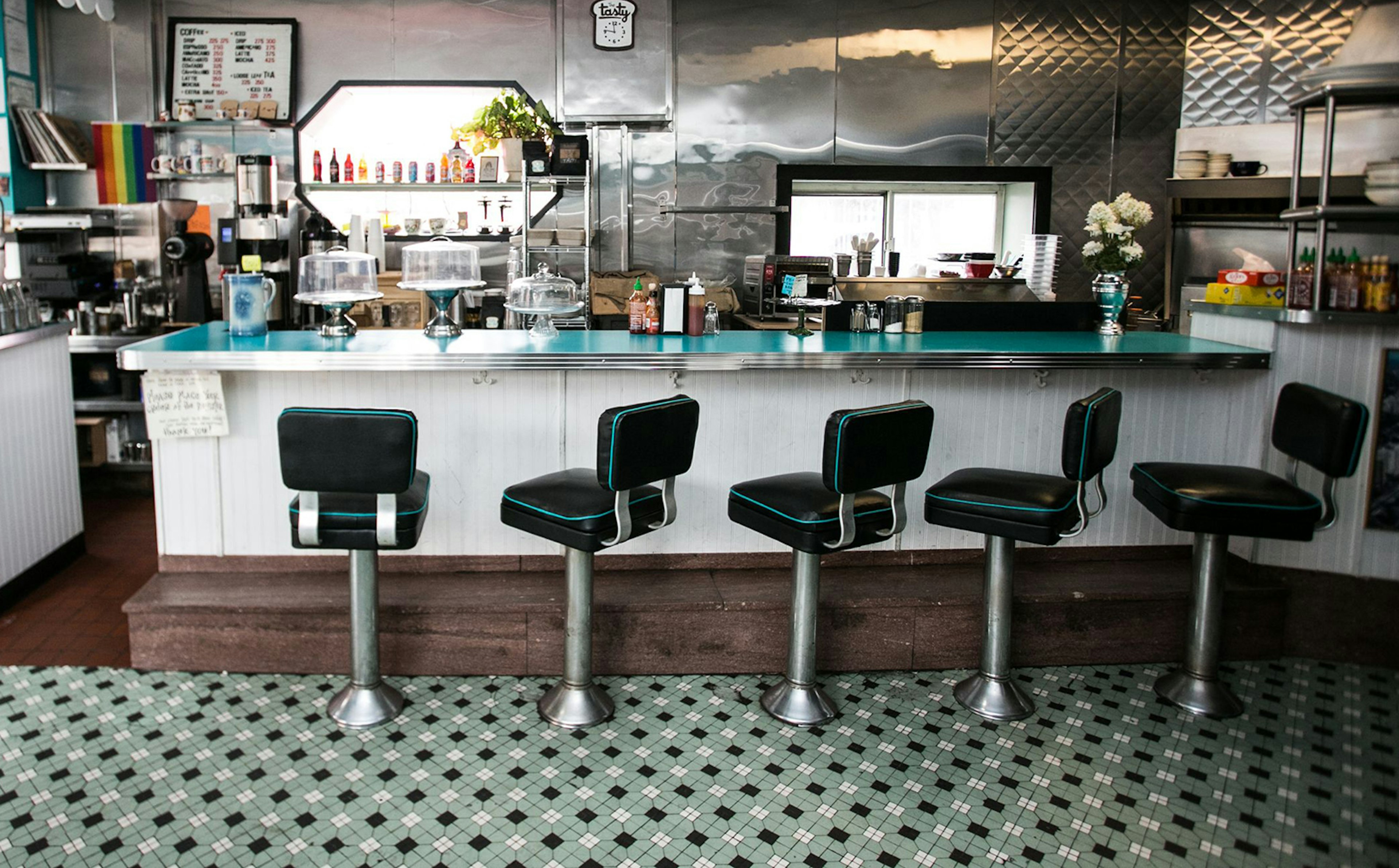 Interior of The Tasty, with five black swivel bar stools on a checkered tile floor in front of a retro, formica counter in a pleasantly cluttered diner