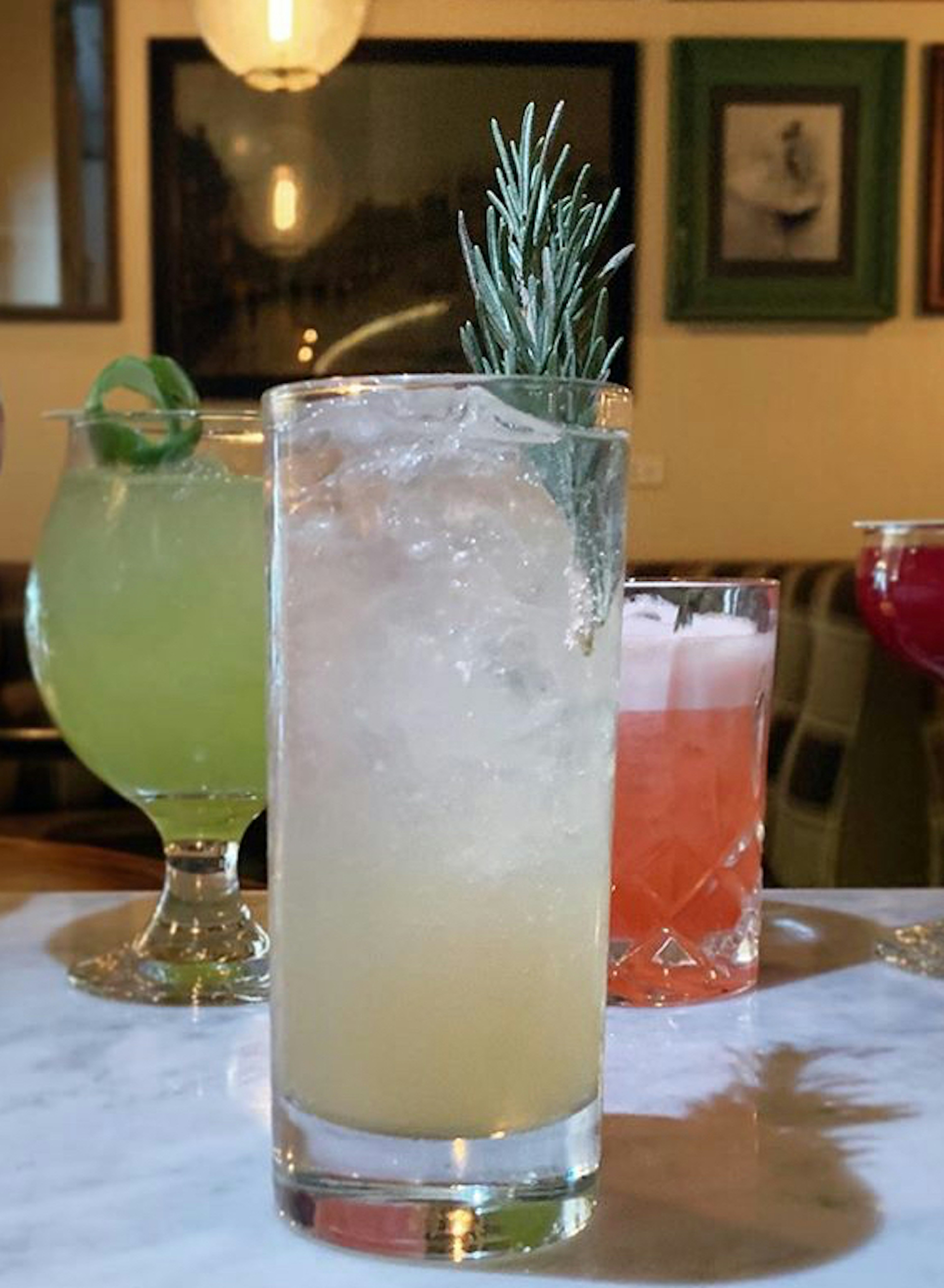 A table has a number of cocktails sitting on it. In the forefront is a white-hued cocktail in a highball glass, garnished with rosemary.