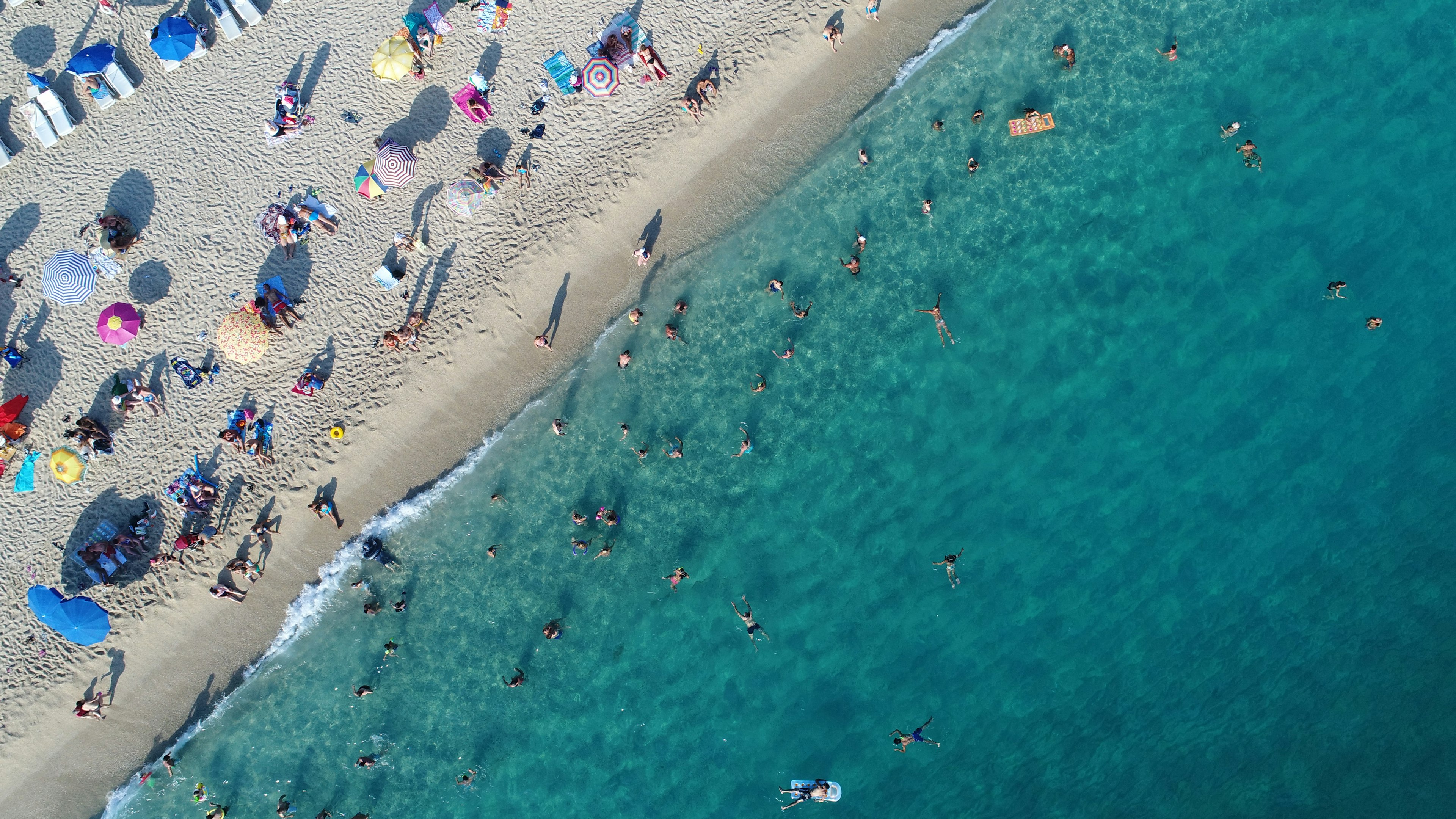 An aerial view of a beach shows colourful umbrellas on the shore and swimmers in the water.