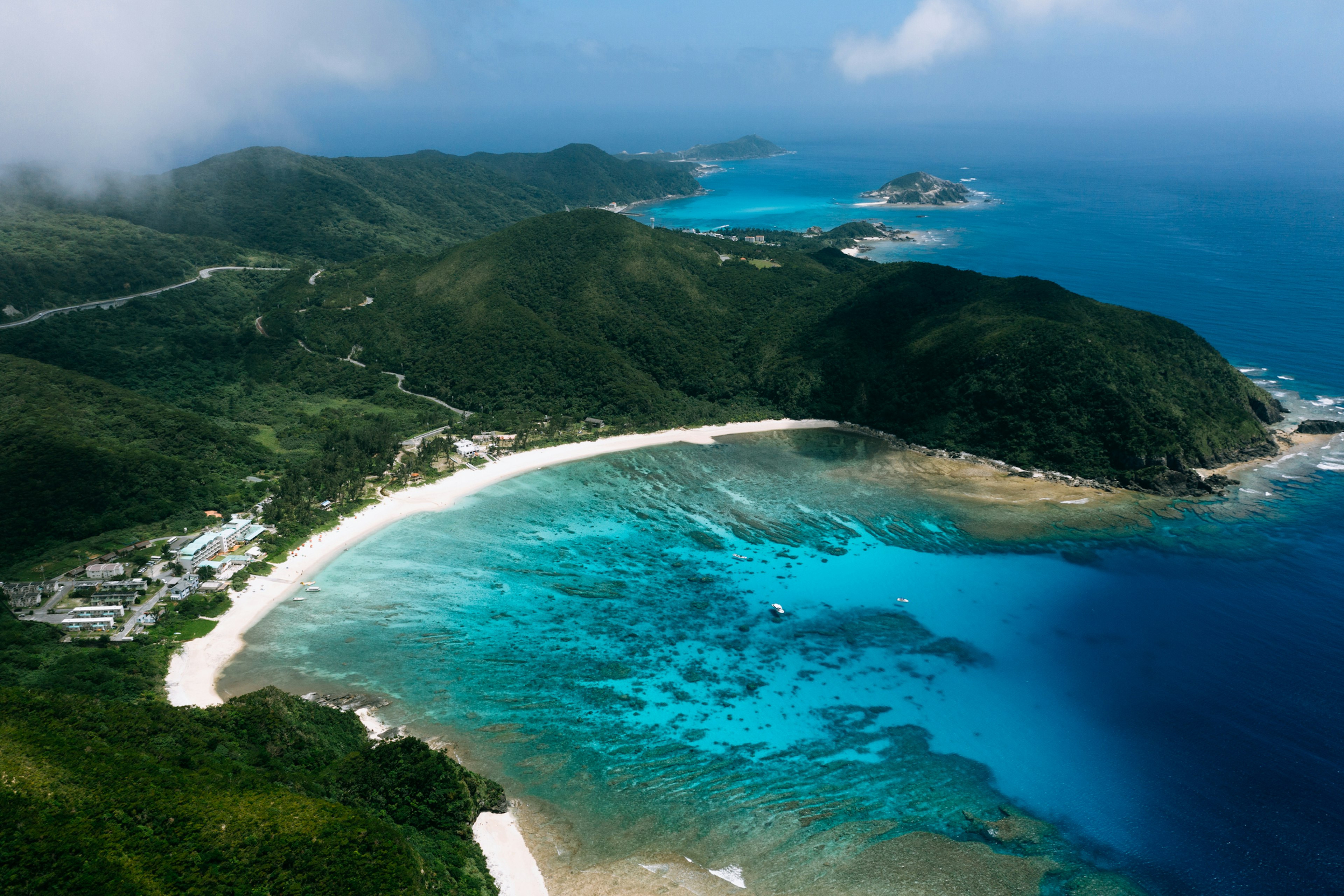 The brilliant blue and teal waters of Keramashoto National Park extend out from a crescent shaped beach, with a thin strip of white sand pressed against the green and black hills of Tokashiki island