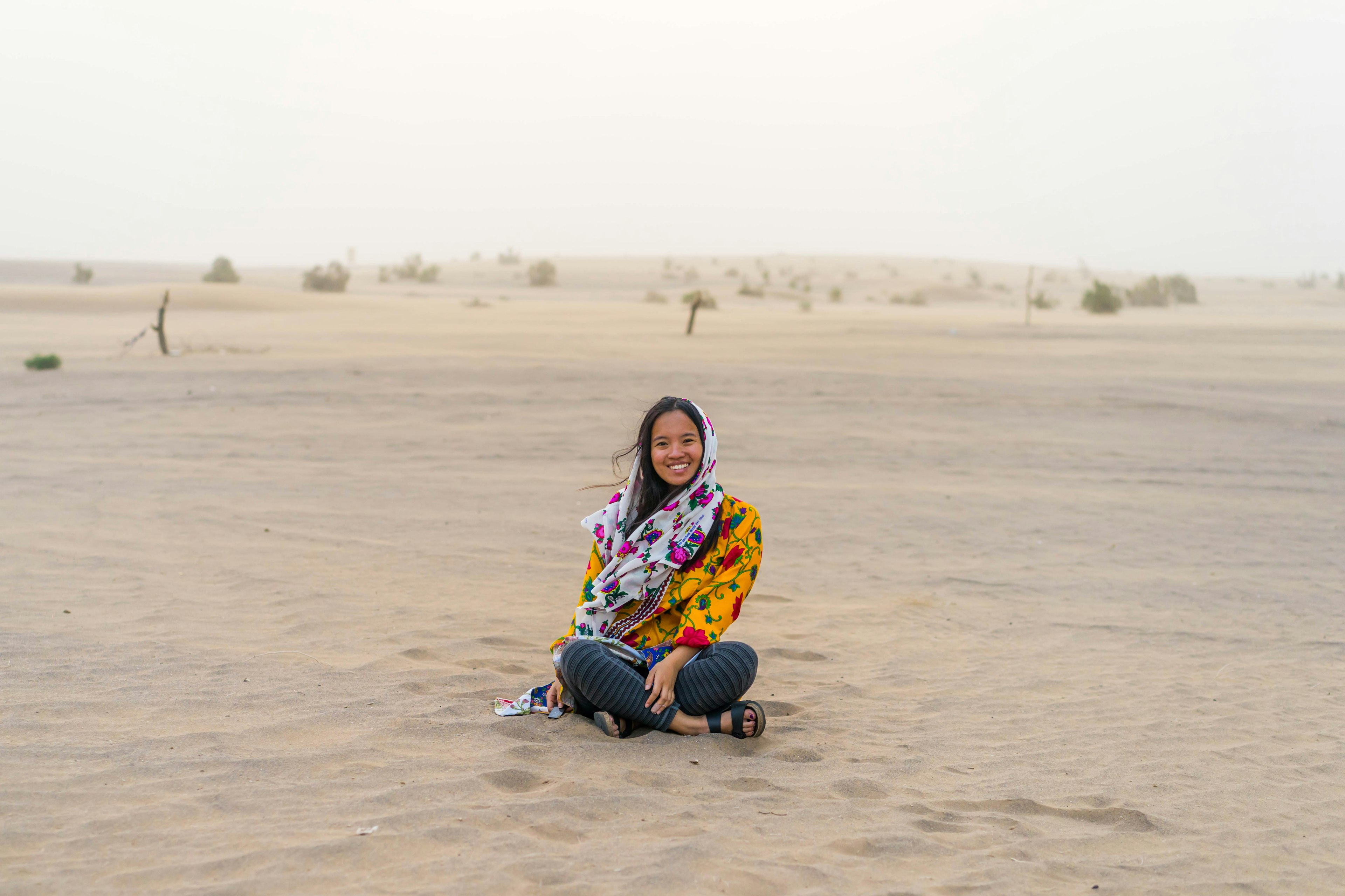 The author sits cross-legged in the desert, smiling, wearing a scarf over her hair