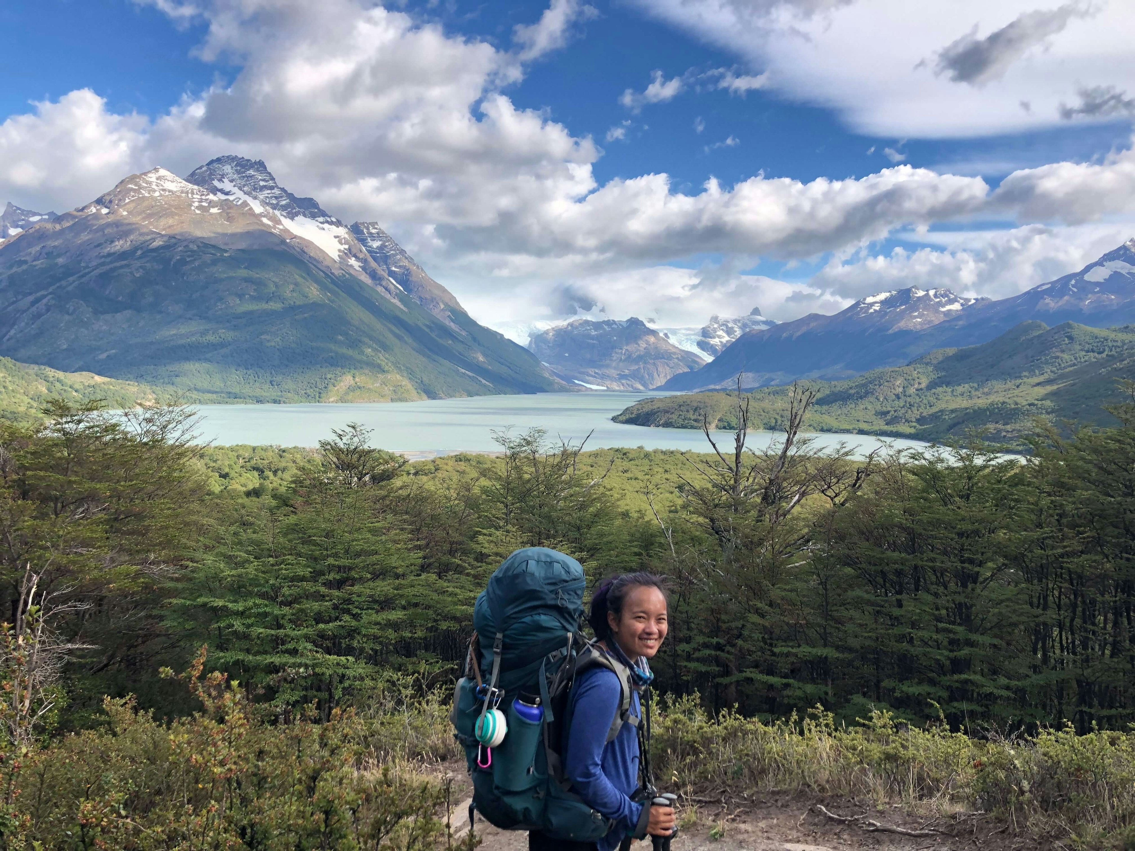 Kay with a backpack and walking pole, with mountains, lake and a valley in the background