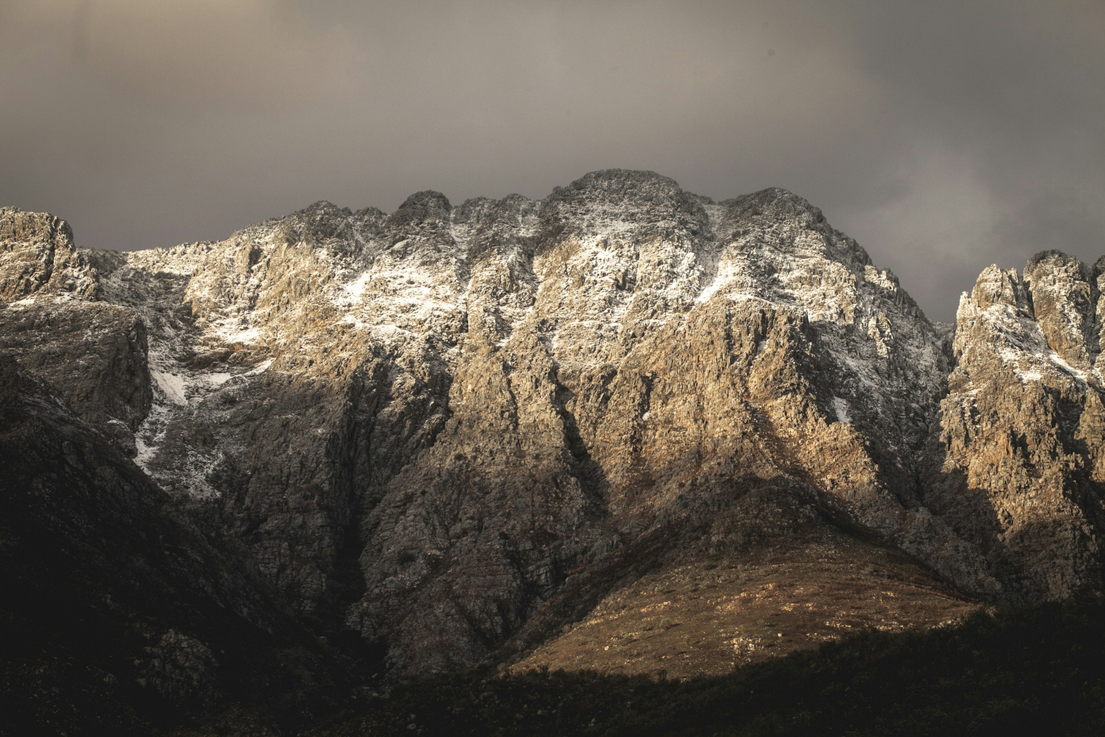 Rounded granite peaks, frosted with snow and lit with a beam of sunlight, stand in front of a dark broody sky