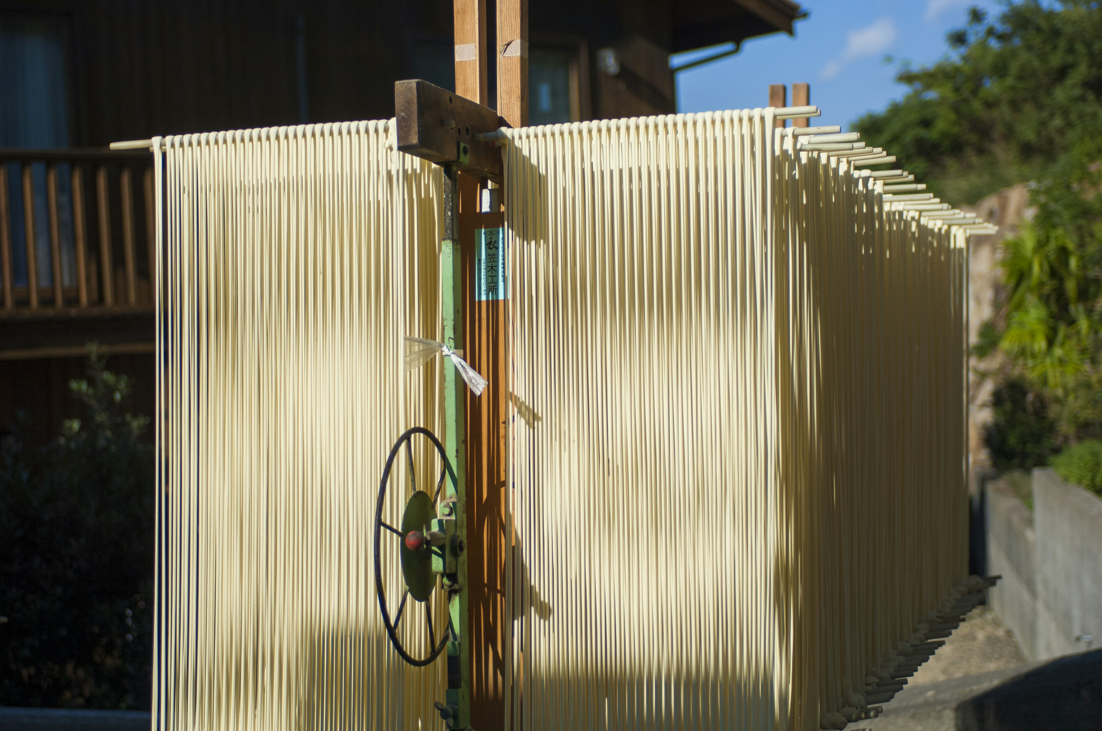 Long strands of shodoshima somen noodles hang from racks outdoors to dry in the sun. The curtains of noodles look almost like a shed made of wood slats they are so long and the racks go back so deep, with a metal wheel in the center of the frame for adjusting the racks