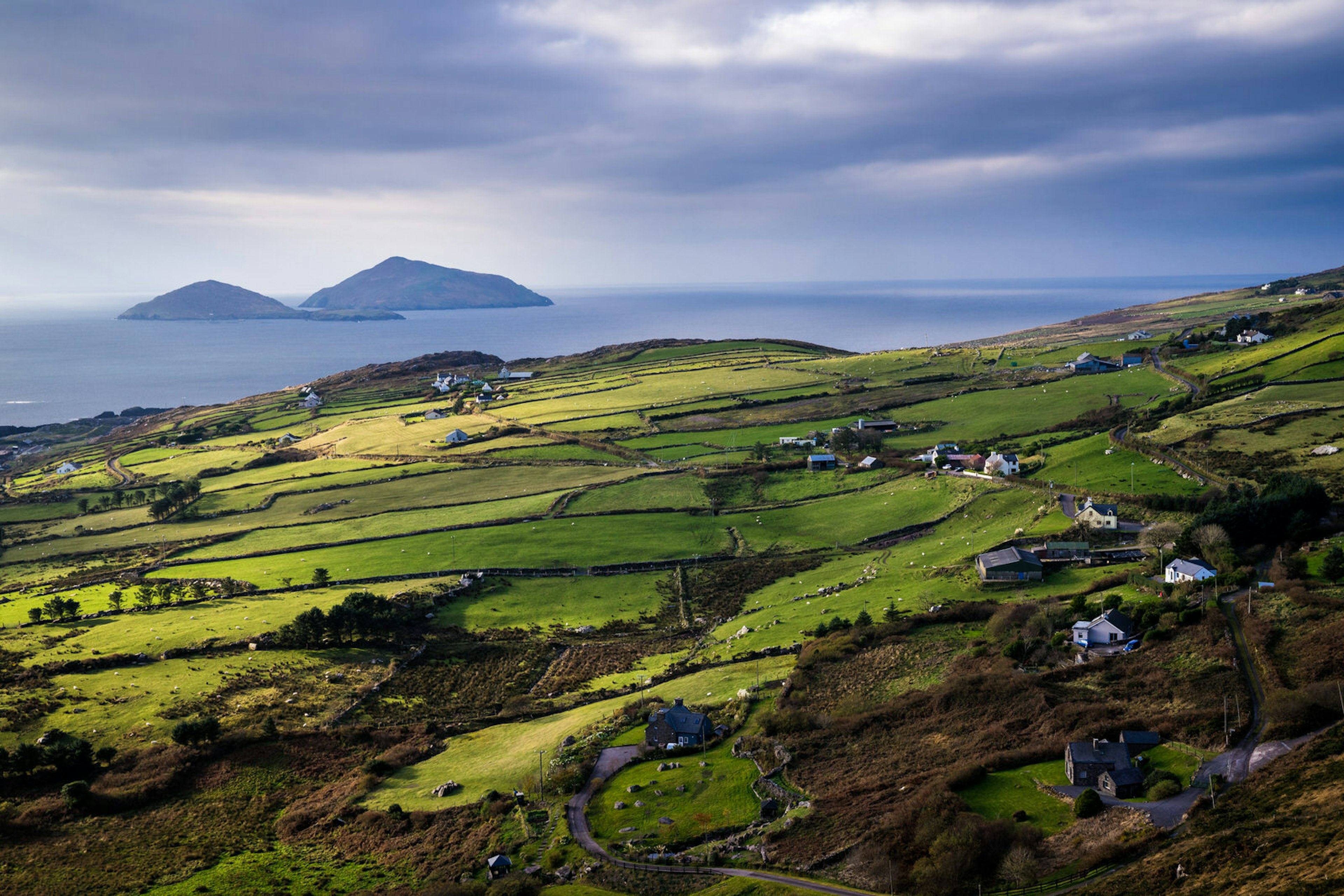 Green rolling fields are in the foreground, with a view of a small rocky island in the distant ocean