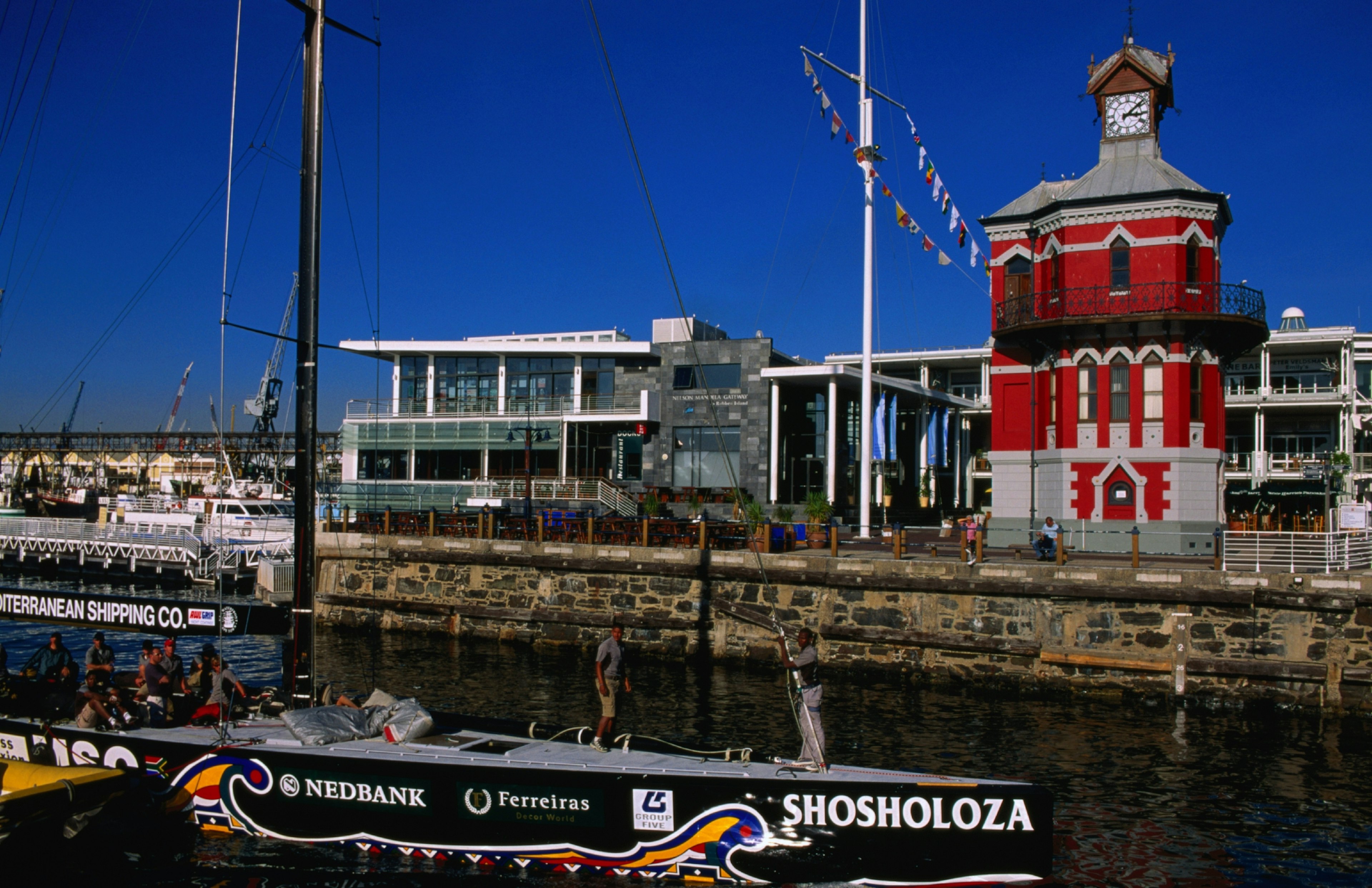 A sailboat sits at a dock, with an historic lighthouse and clocktower in the background; the sky is cobalt blue.