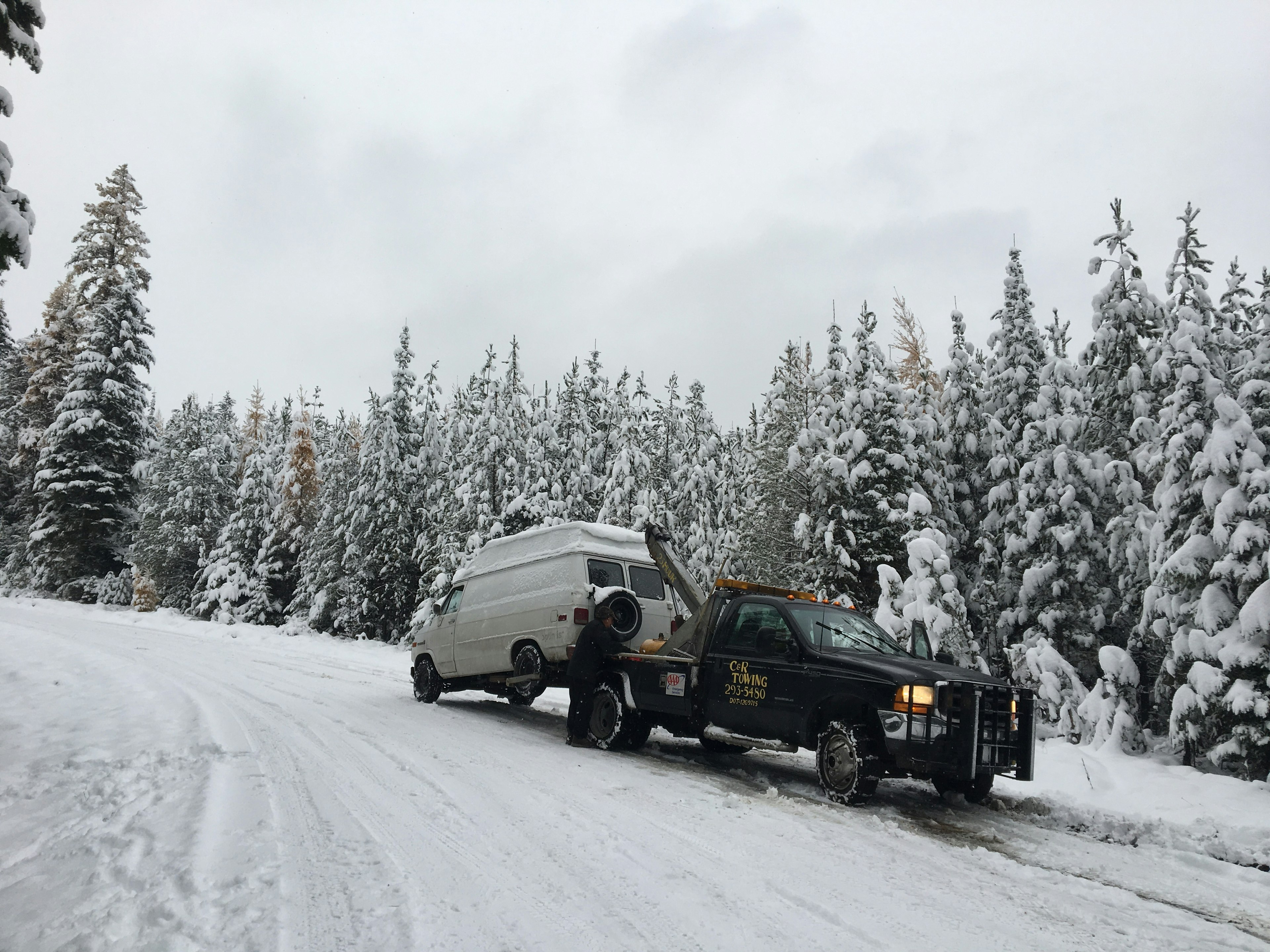 A white GMC van is being towed by a black wrecker on a snowy road in Montana. Spruce and pine trees in the background at blanketed with snow, as is the forest service road where the van broke down.