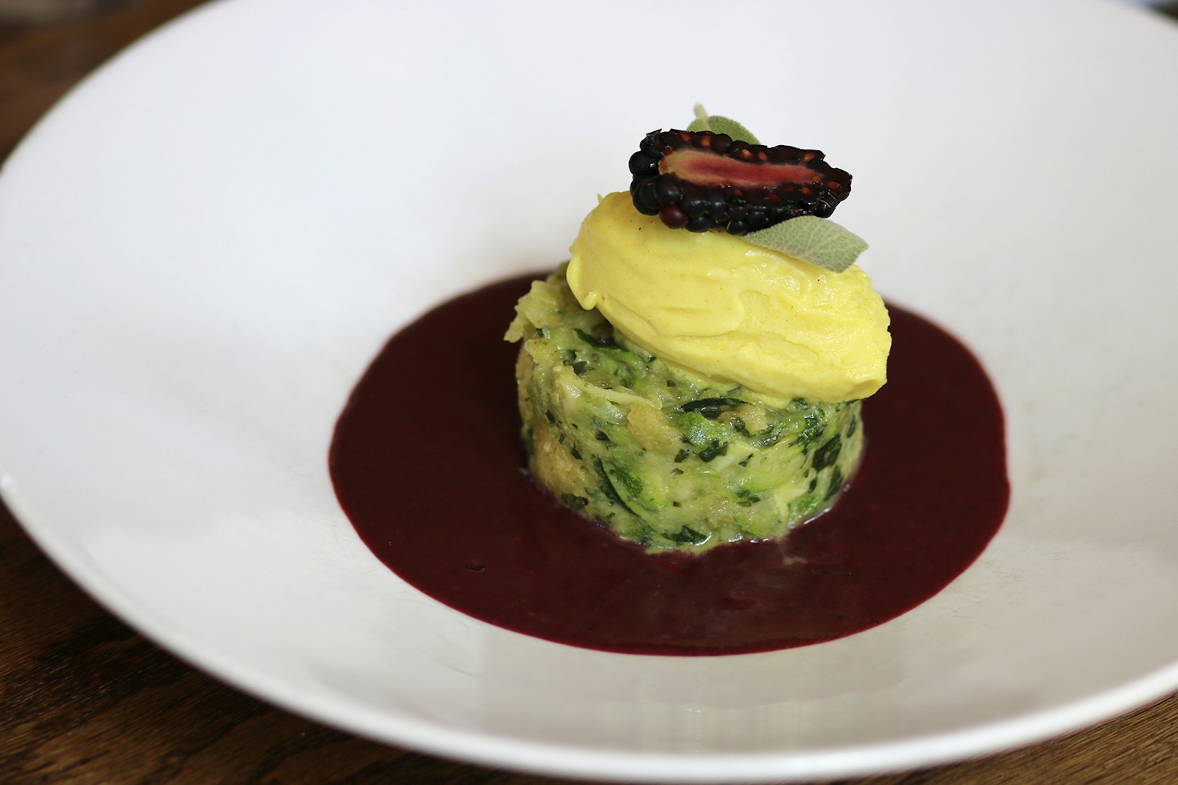 Close-up of pale green zucchini bread pudding, topped by a pale yellow vegan bread and half a blackberry and surrounded by a dark, sweet blackberry sauce