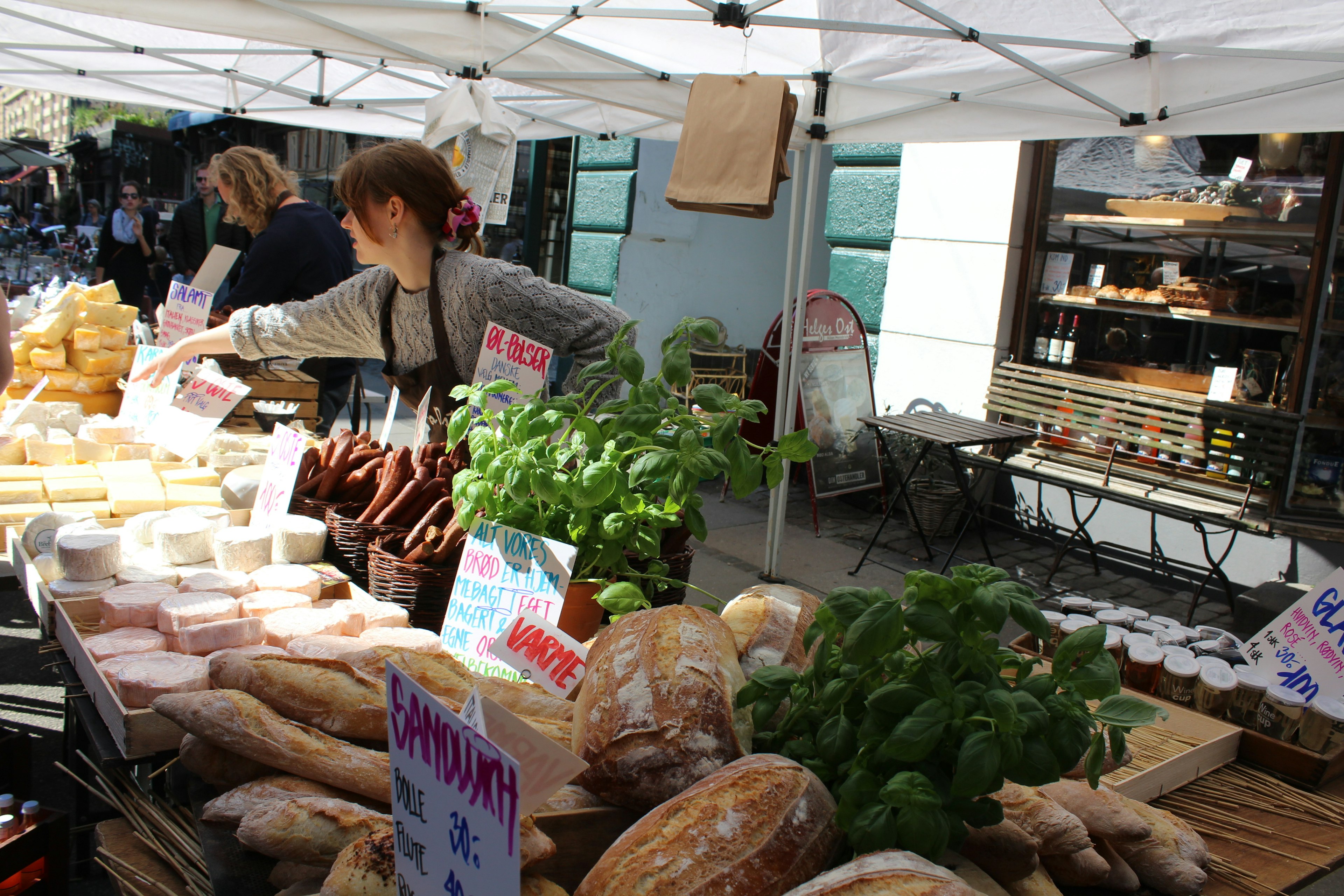 A close up shot of a stall at a street market with a white gazebo roof. The stall is selling basil, cheese and crusty bread. A woman in a woolly jumper and brown apron is reaching over to serve a customer.