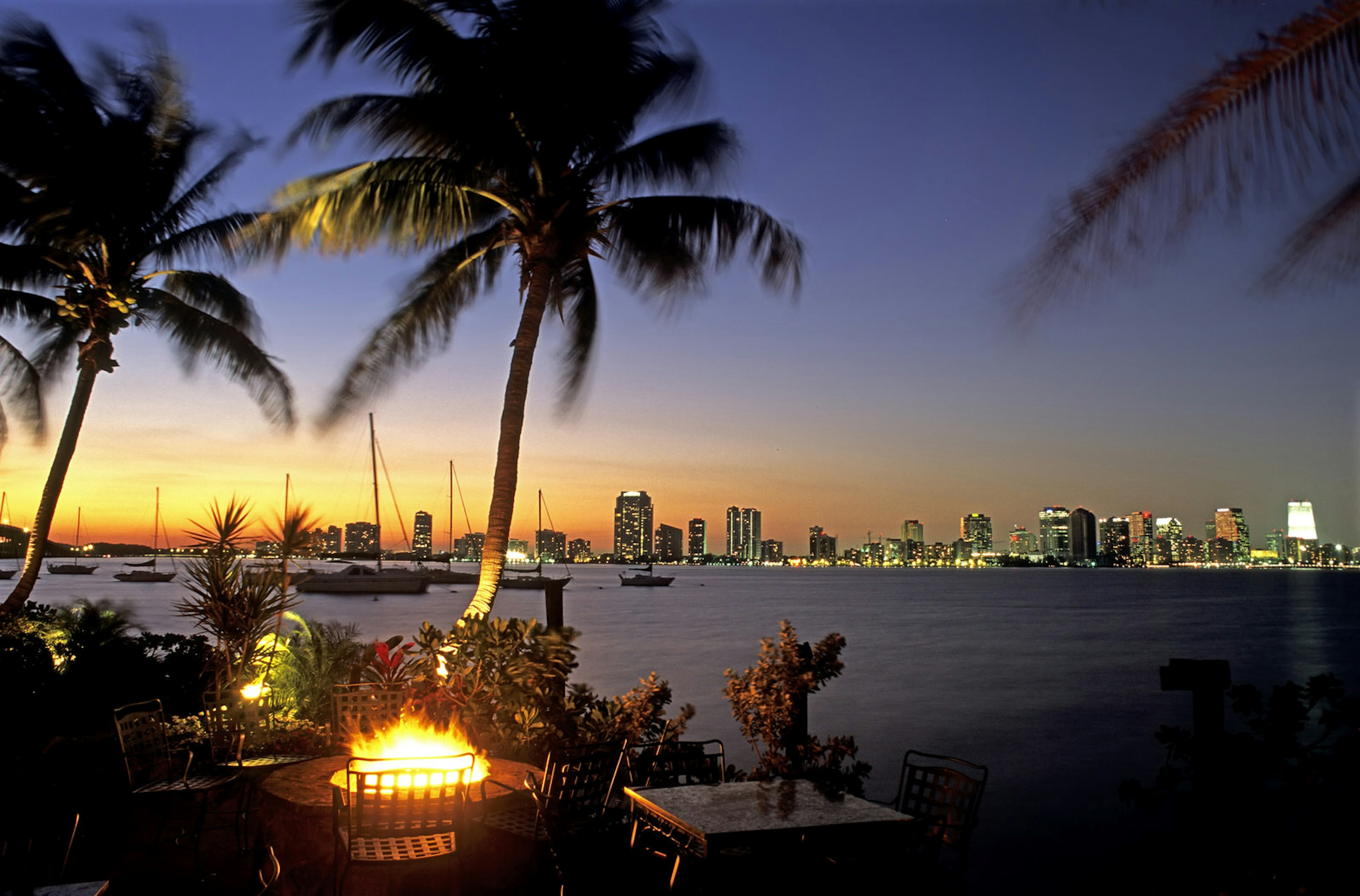 A long-distance view of the Miami skyline is seen over a dark bay at sunset. In the foreground is an outdoor bar area with palm trees and a fire pit © Louis Davilla / Getty Images