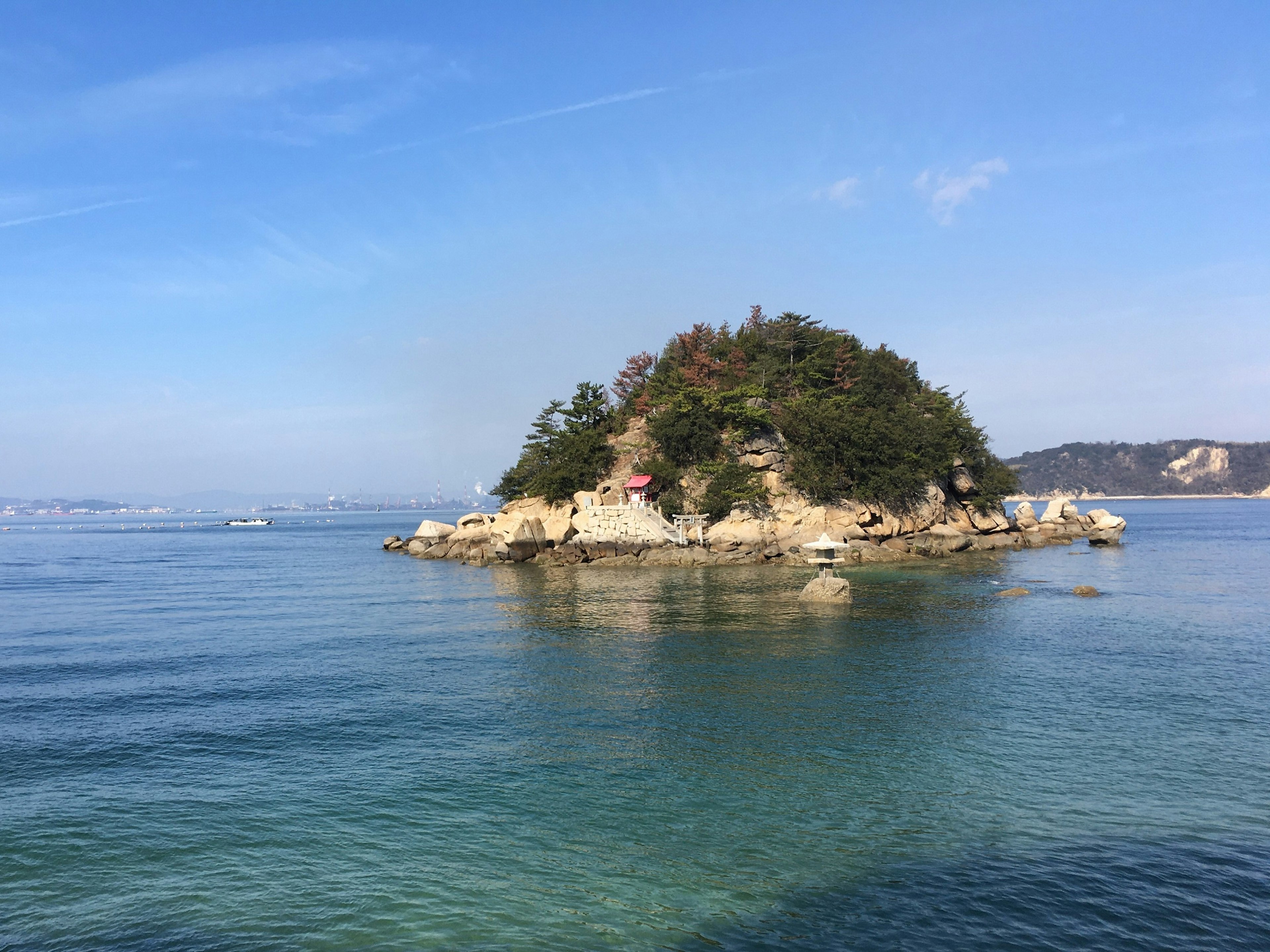 Very still sea, with a small rocky island covered in trees. A small shrine with a red roof is on the edge of the island.