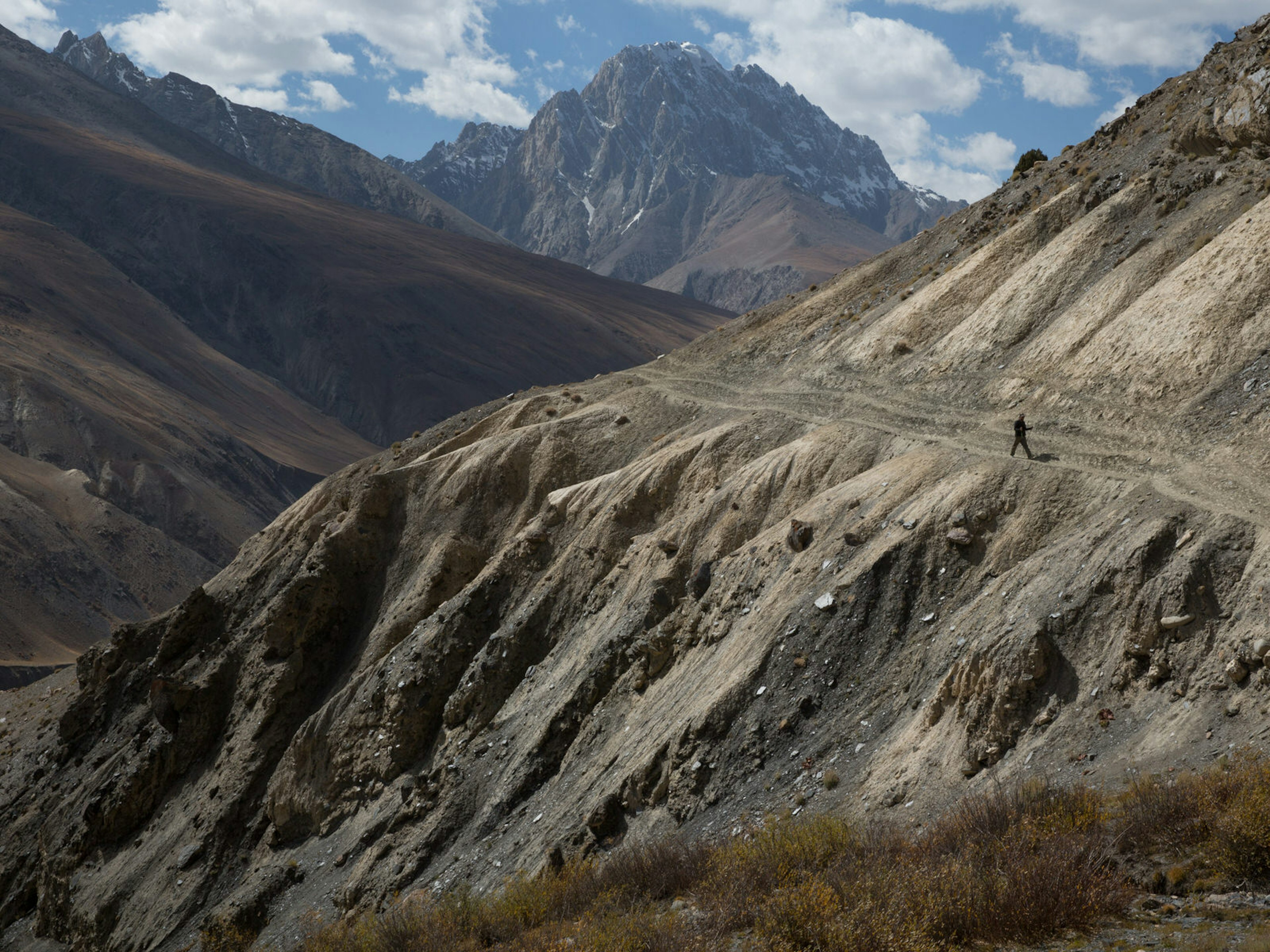 A lone walker treks along a steep mountainside path with tall peaks in the background