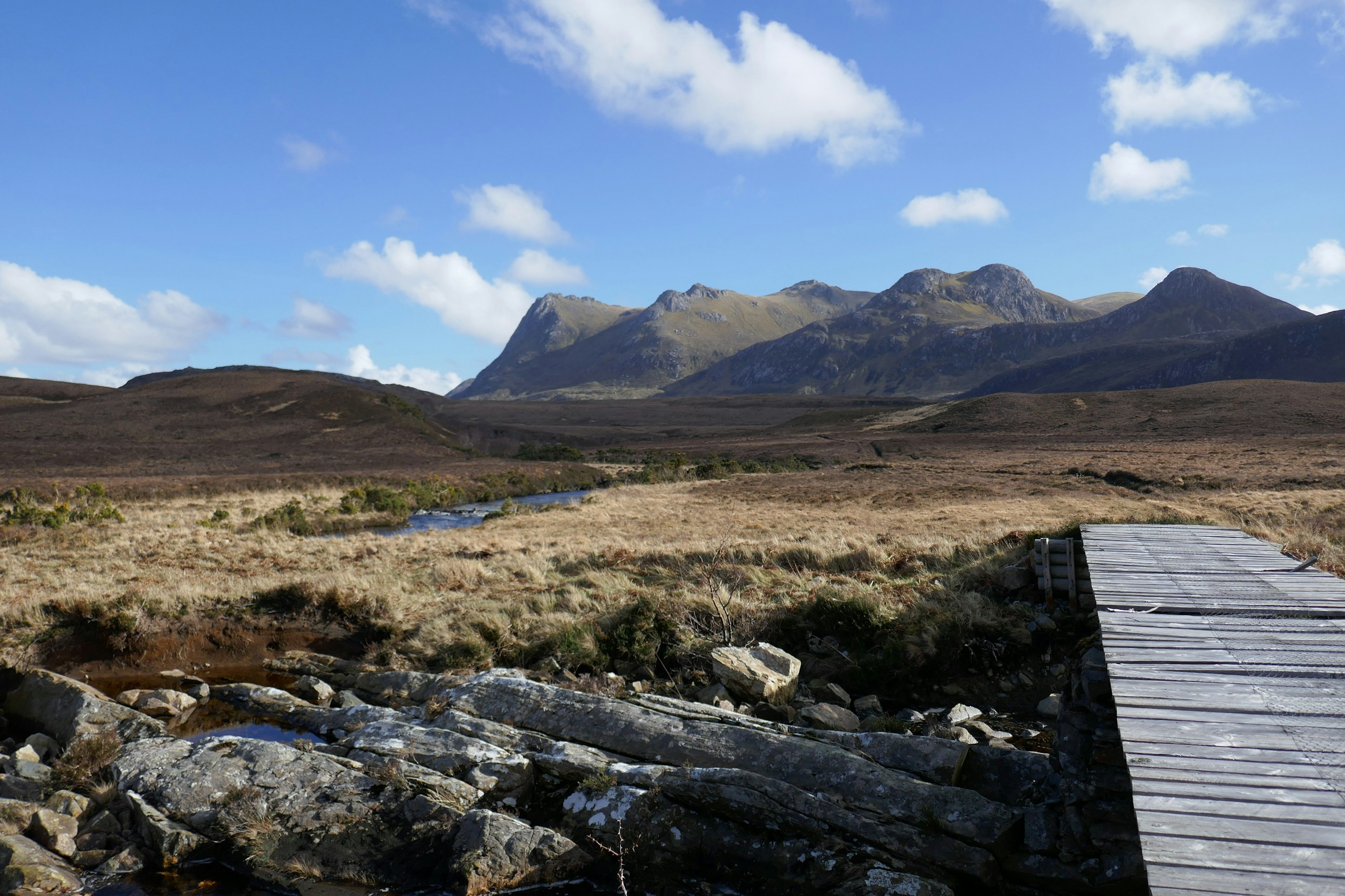 A boardwalk crosses a ditch leading into wild grassland. Several large hills are in the distant