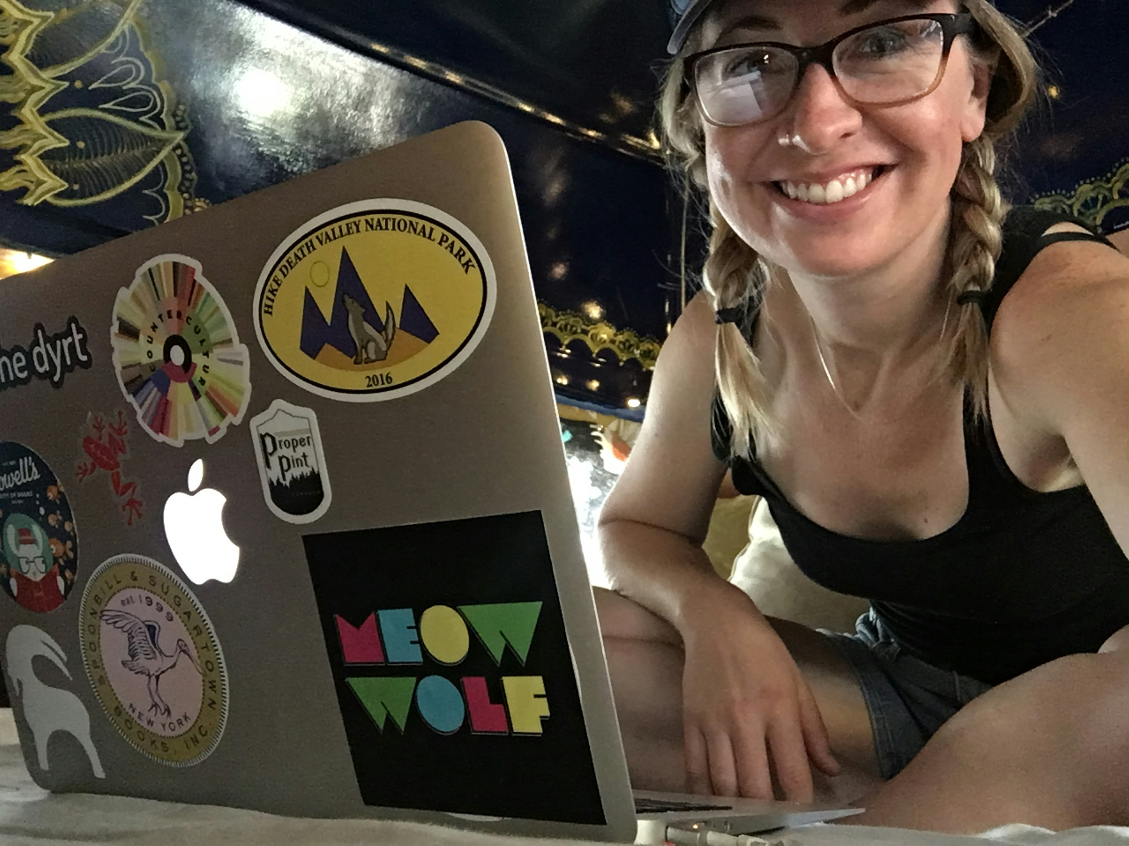 A thirty-something year old woman sits with her legs crossed in shorts and a black tank top in a van, her light brown hair braided in two plaits and brown and black glasses framing her face. Her laptop is in the foreground, covered in colorful stickers.