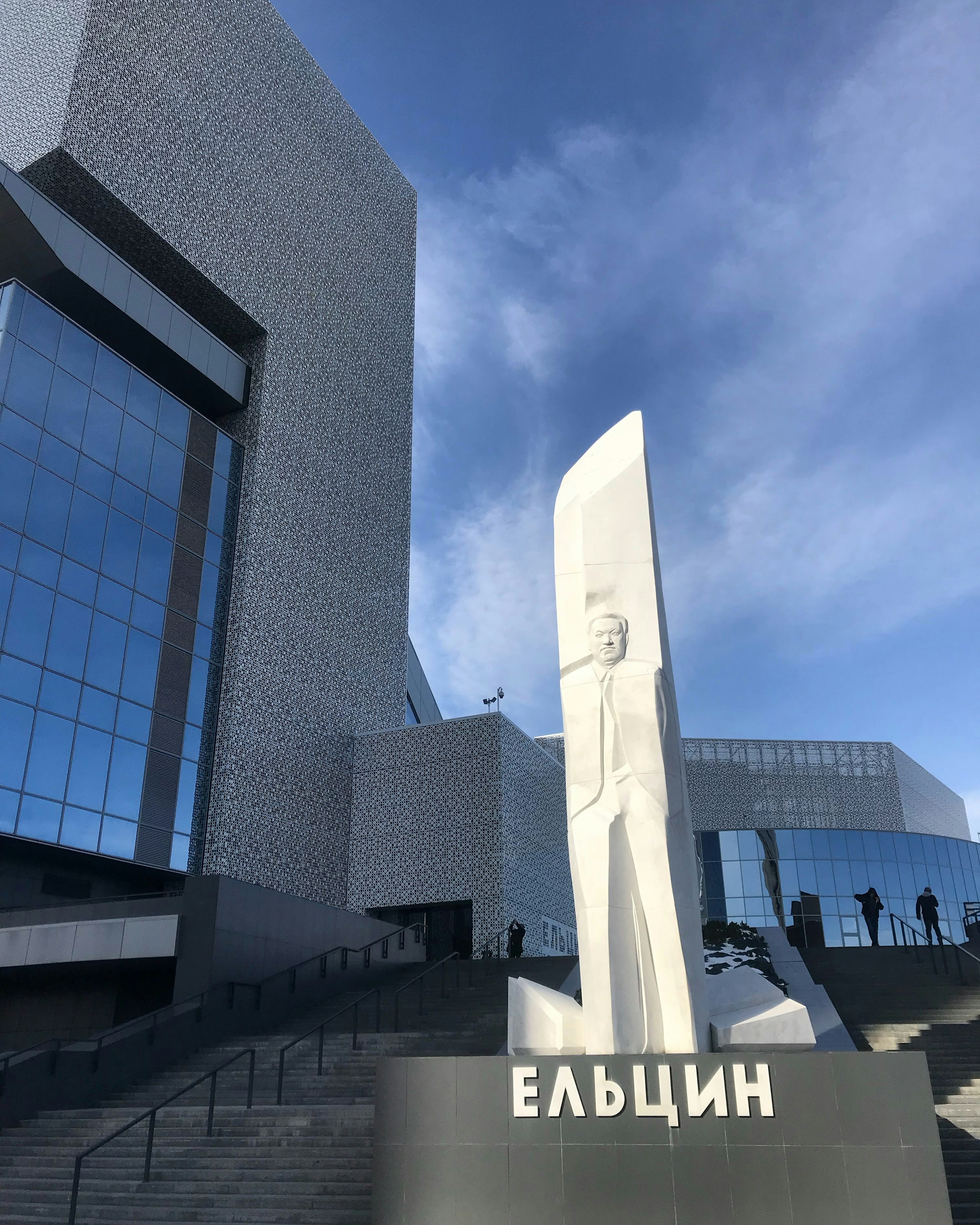 A tall white relief sculpture depicting a man stands in between two flights of steps. A large angular modern grey building is in the background.