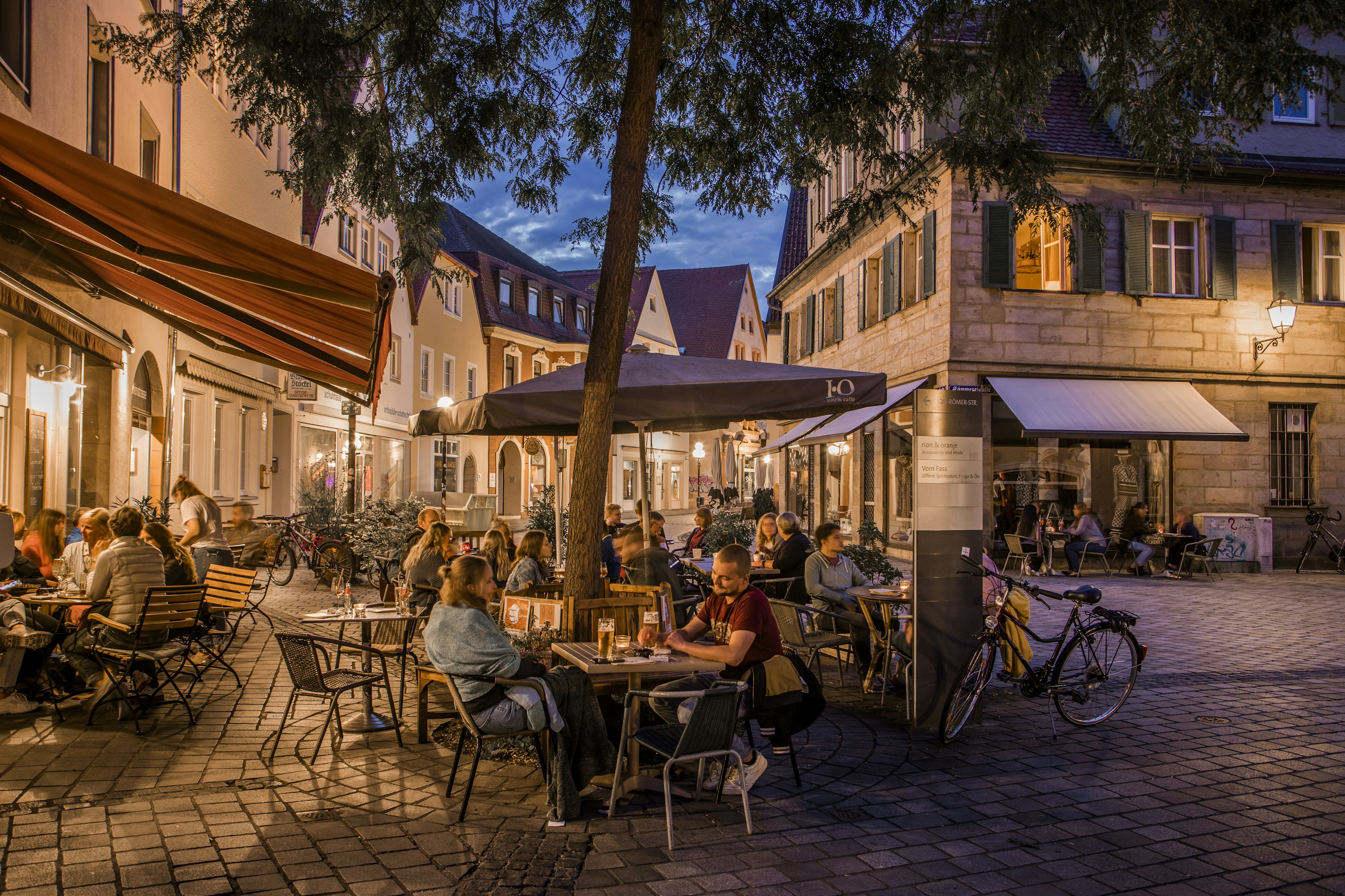 A nighttime scene shows many people sitting at outdoor tables at a cafe in Bayreuth, Germany, with a cobblestoned square and bicycles around.