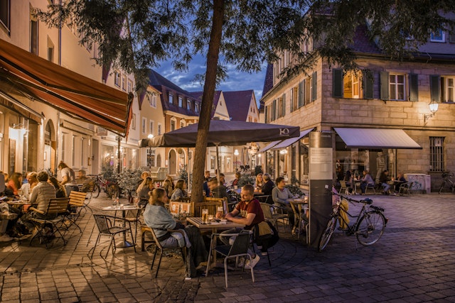 A nighttime scene shows many people sitting at outdoor tables at a cafe in Bayreuth, Germany, with a cobblestoned square and bicycles around.