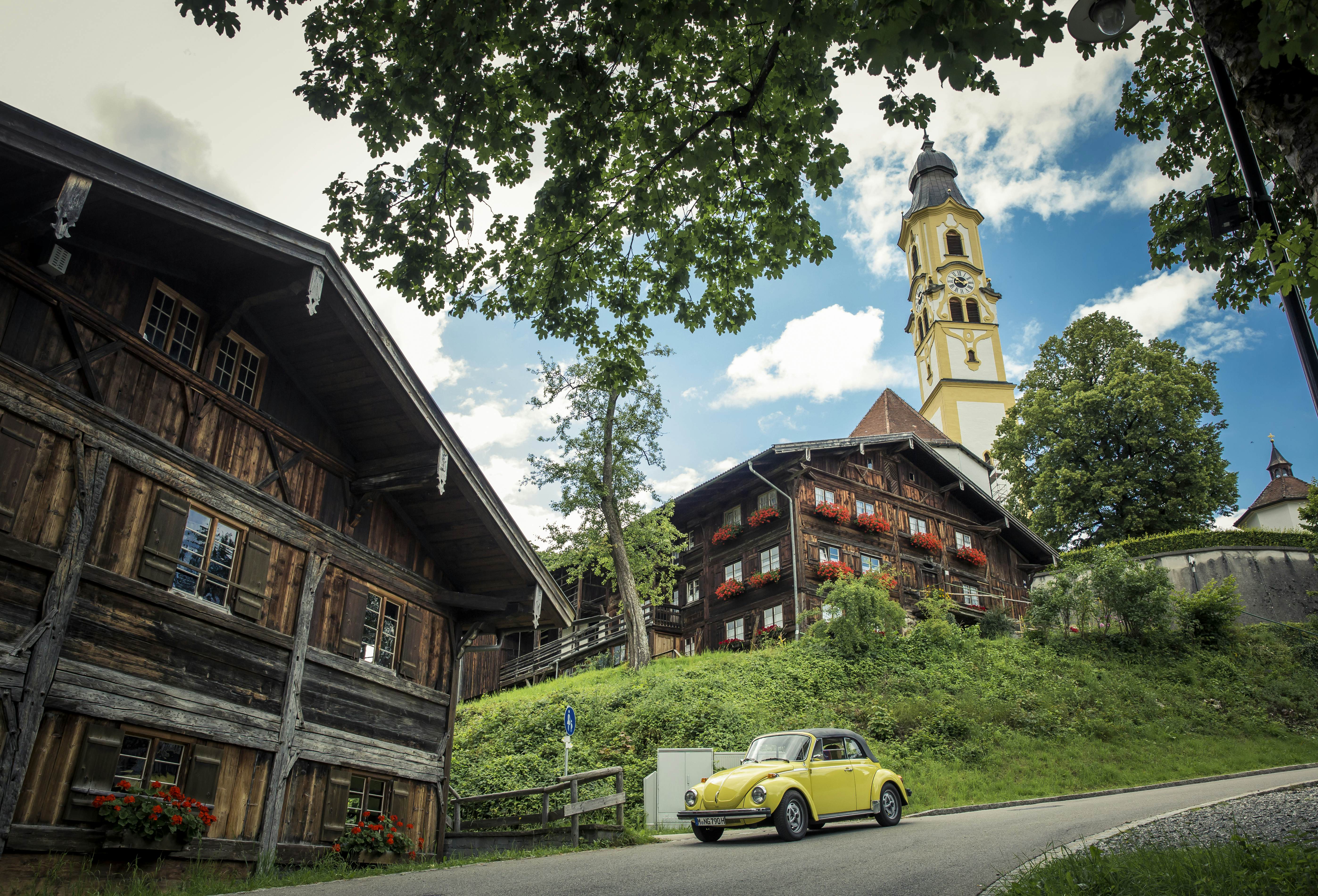 A yellow Volkswagen Beetle passes by Alpine chateaus and a building with a yellow tower on the German Alpine Road in Bavaria