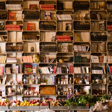 A wall of compartmentalised bookshelves is filled with books; some shelves also have wine bottles and fresh squashes. A table of inviting fruit and vegetables sits underneath the books.
