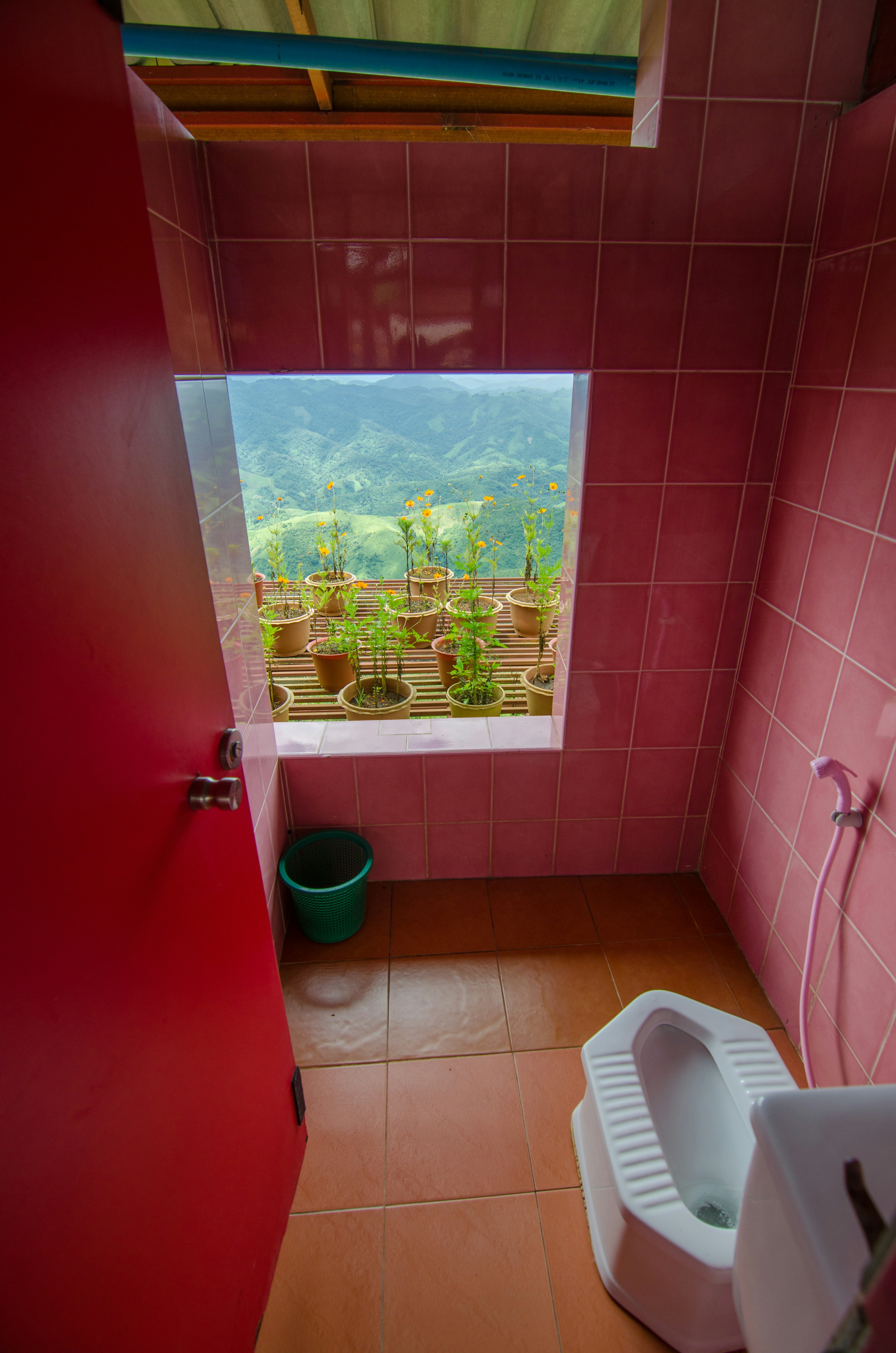 Toilet at a road-side rest spot in Laos. The bathroom has an open wall overlooking a beautiful valley and a ledge with flower pots on it.